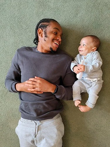 **Alt text:** A smiling adult and baby lie side by side on a soft green rug, looking at each other.