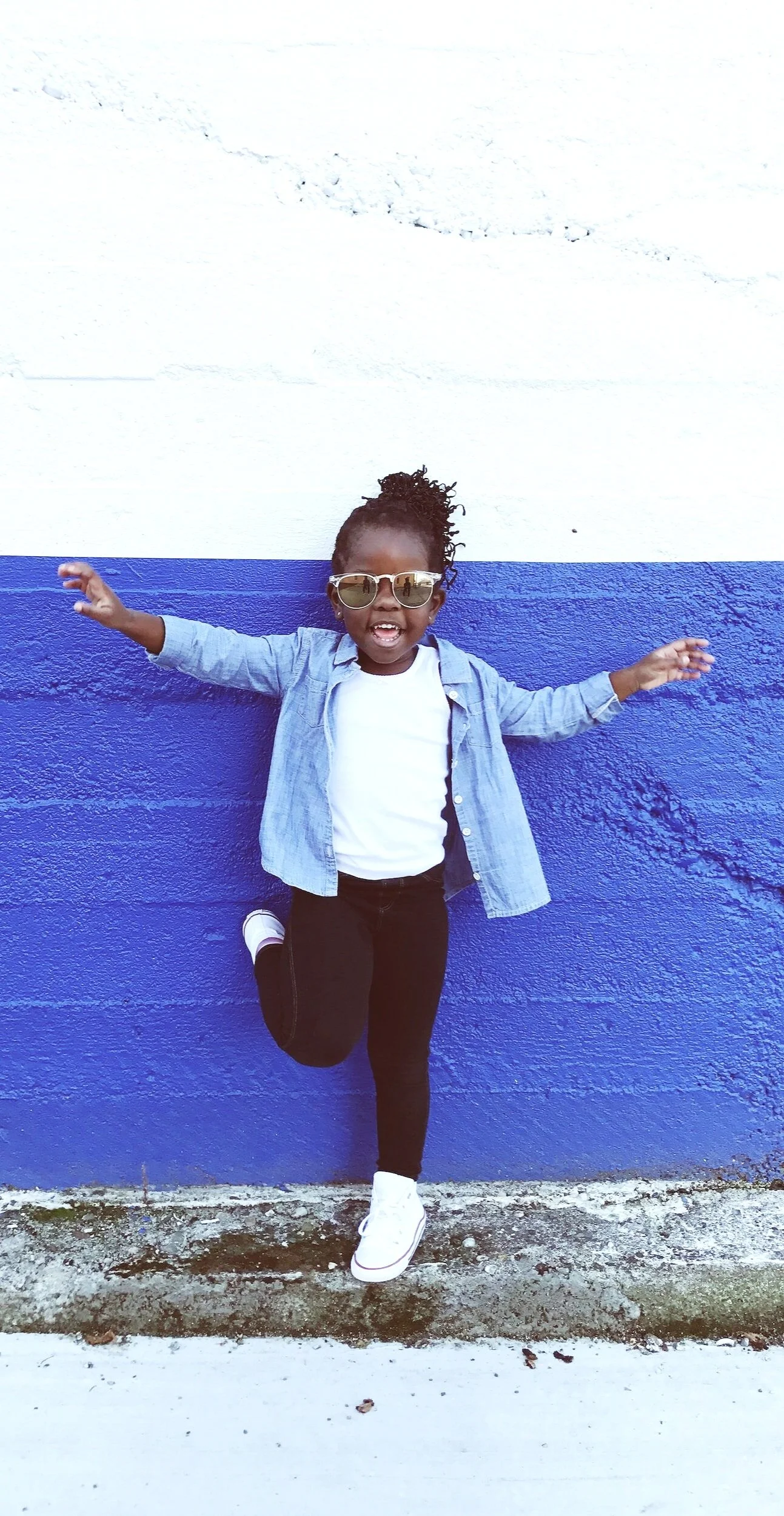 A young girl in sunglasses, a white shirt, and a denim jacket standing against a two-tone wall, smiling and posing with arms outstretched and one leg lifted.
