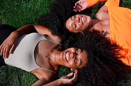 Two smiling women with long curly hair lie on green grass. The photo is taken from above in bright sunlight, creating a relaxed, joyful atmosphere.