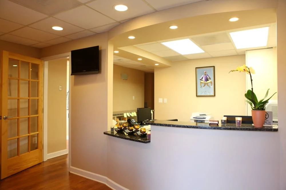 Reception area with a curved counters, a yellow orchid in a pot, several candles, a computer, a framed picture, and a wall-mounted TV. Wooden door with glass panes to the left, ceiling lights, and hardwood flooring.
