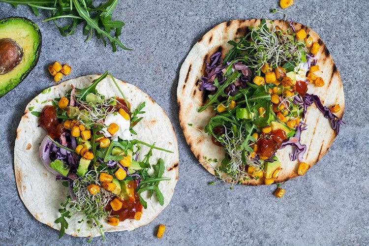 Two open-face tacos with lettuce, corn, purple cabbage, microgreens, and a drizzle of sauce on a gray surface, with a sliced avocado and some corn kernels nearby.