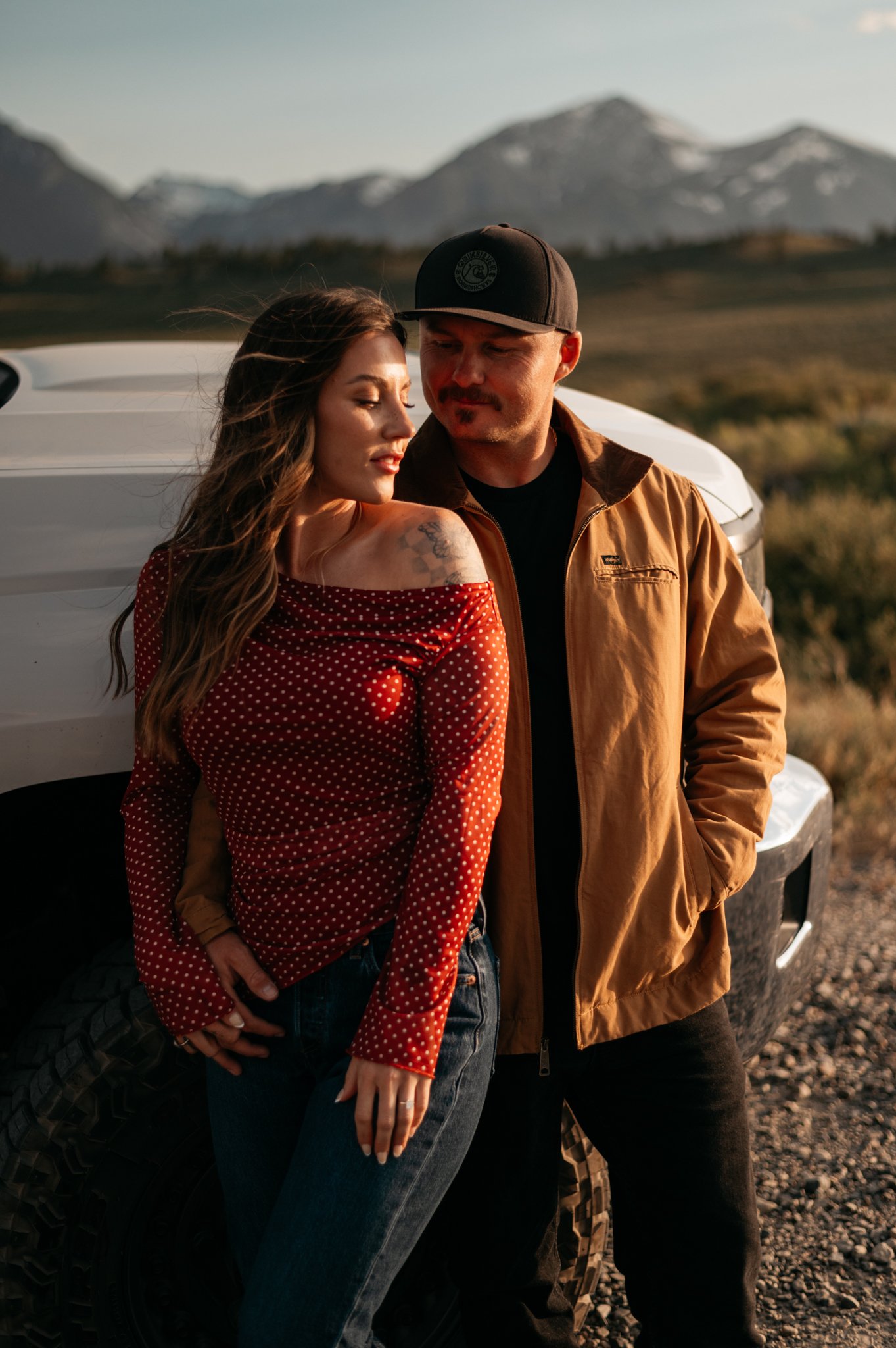 A young woman with long brown hair, wearing a red polka-dot off-the-shoulder top, and a man with a beard, wearing a black cap and brown jacket, leaning against a white 4x4 truck outdoors with the eastern sierra mountains in the background. The man ju