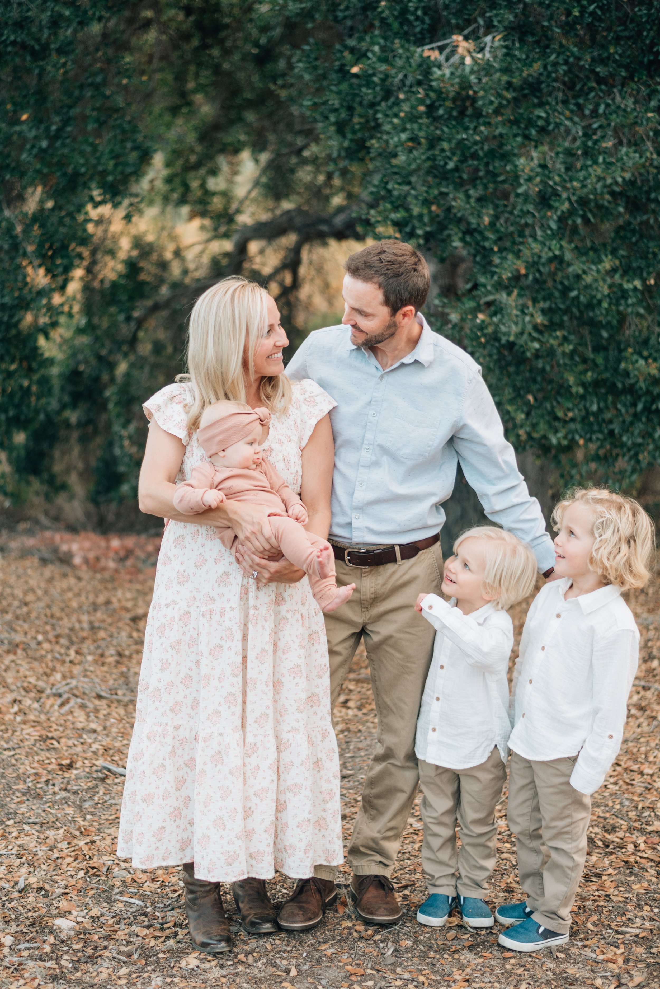 A family of five standing outdoors, with a woman holding a baby girl and a man and two young boys standing next to her, all smiling at each other.