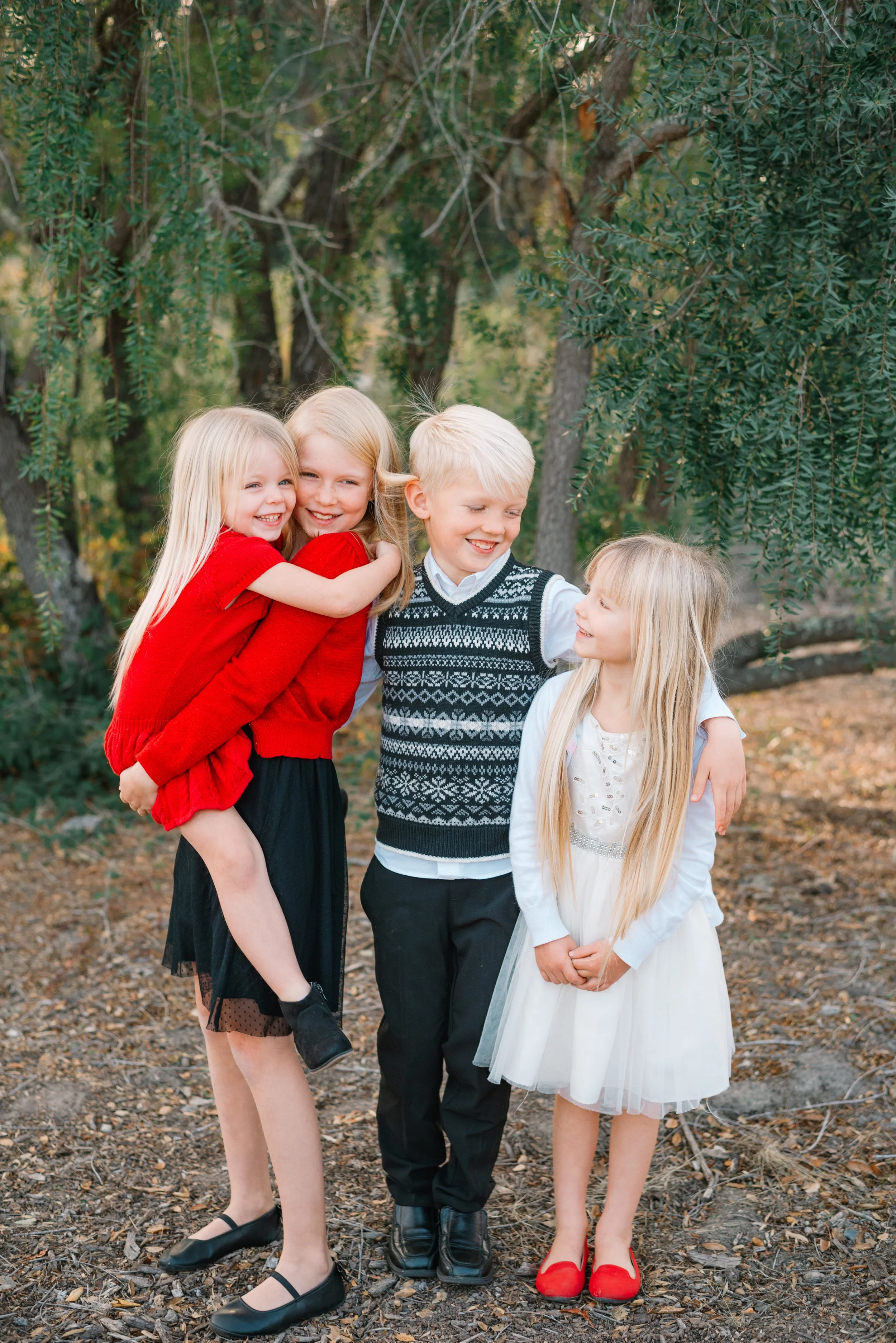 Group of four young children, two girls and two boys, sharing a funny moment outdoors in a wooded area, dressed in festive holiday attire.