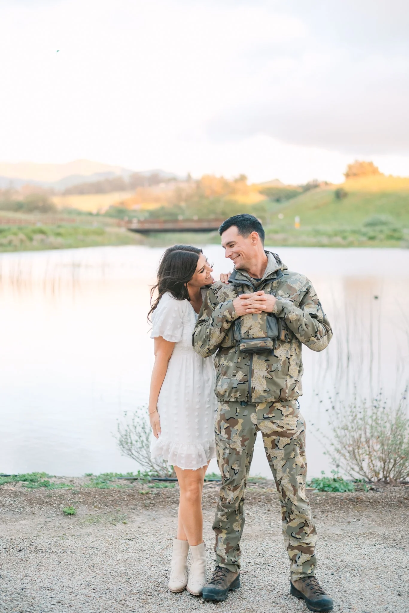 A woman and a man, the man in military camouflage uniform, are standing by a lake, smiling and looking at each other affectionately during sunset.