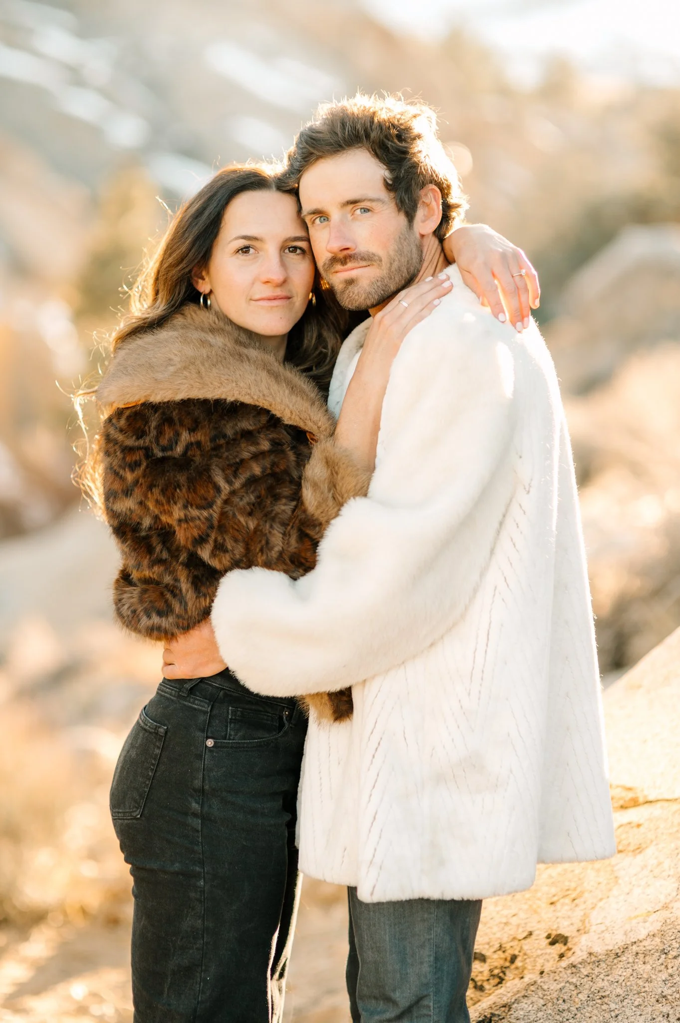 A couple embracing outdoors during golden hour, both wearing cozy winter clothing, with a blurred natural background.