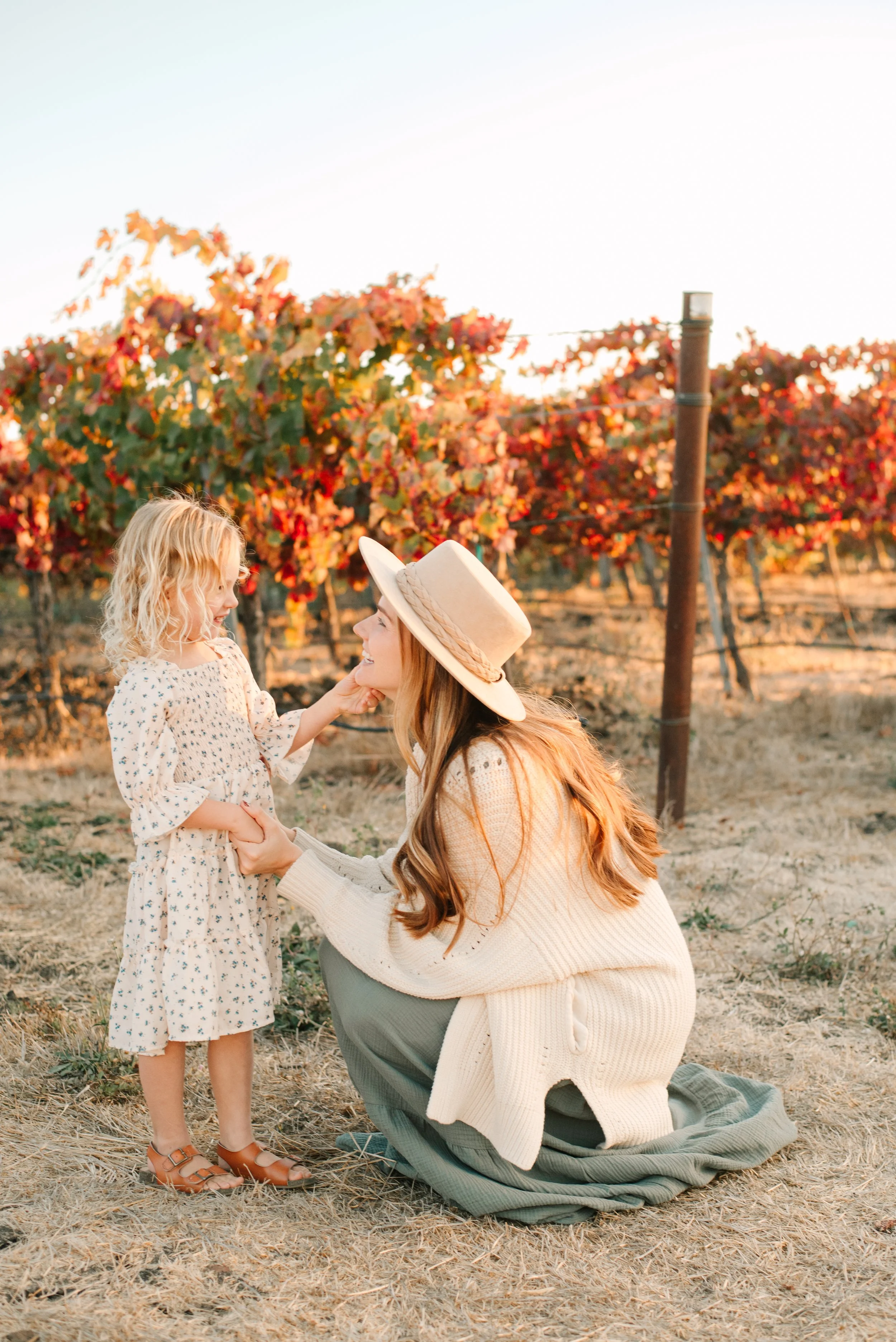 A woman in a wide-brimmed hat kneeling on the ground, holding a young girl’s hands, with a vineyard with red and orange leaves in the background.