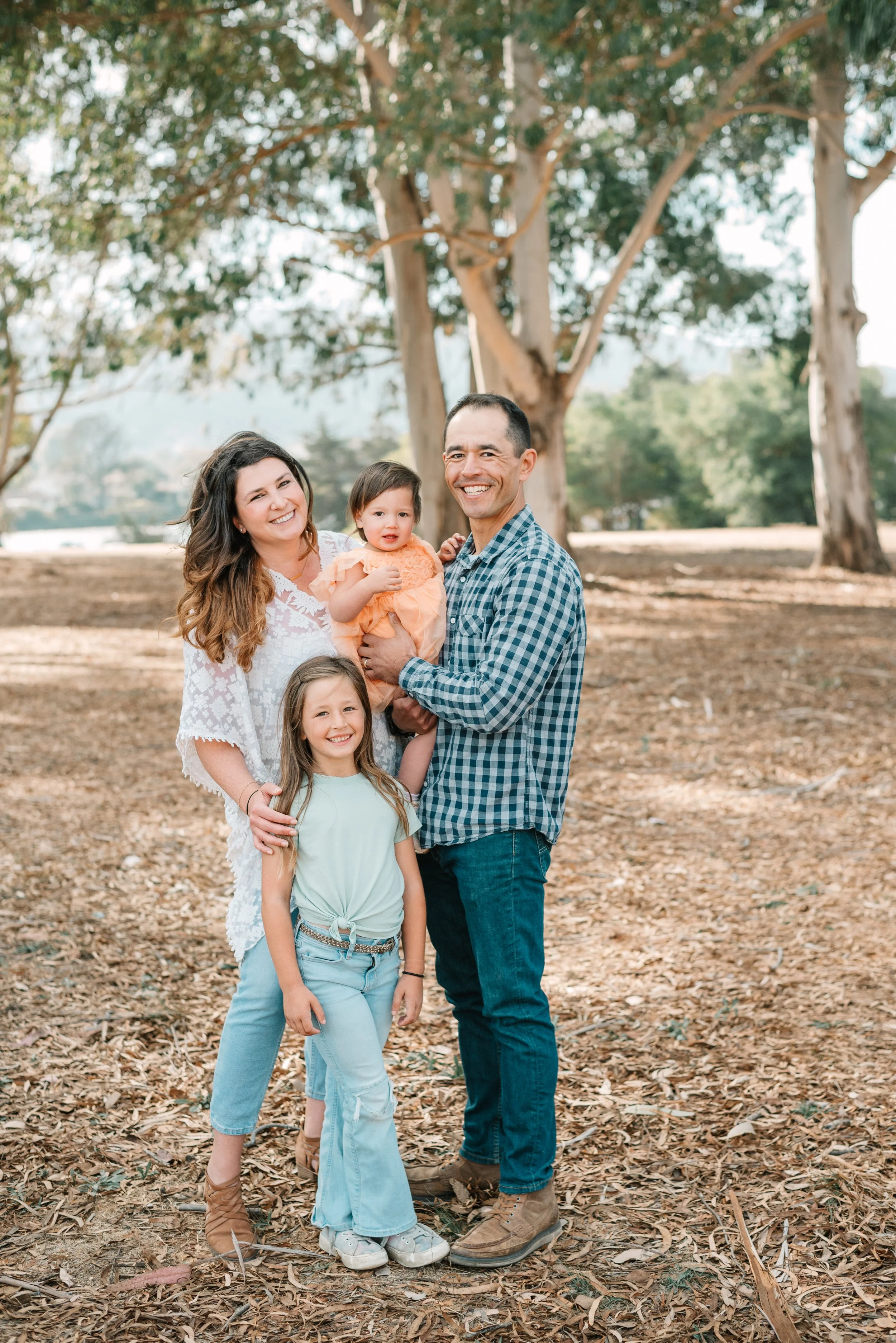 A smiling family of five standing outdoors on a leaf-covered ground with large trees in the background, including two young girls, a woman, a man, and a baby girl.
