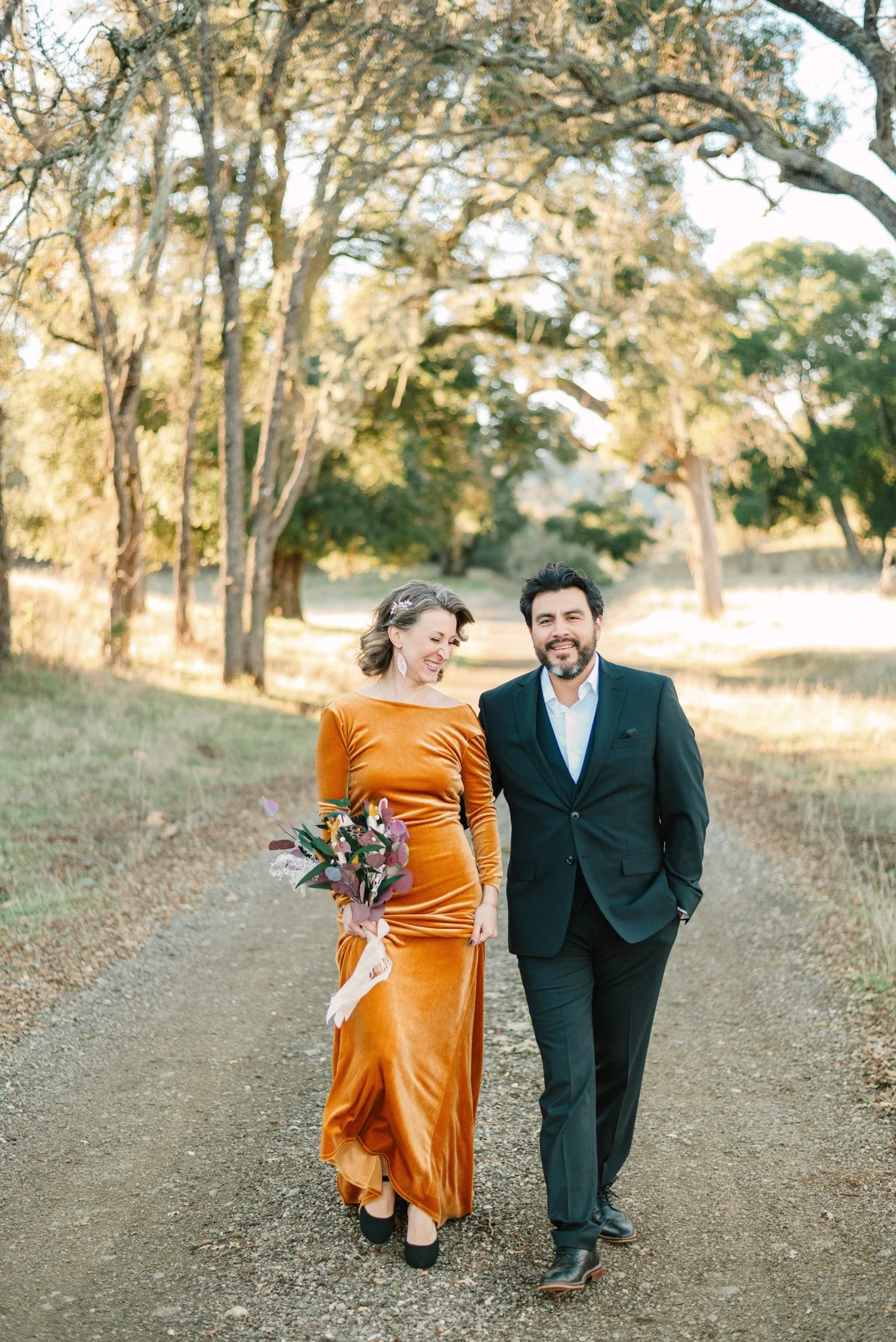 A woman in a long, orange velvet dress holding a bouquet of flowers walks with a man in a black suit and white shirt along a dirt path in a forested area during daylight.