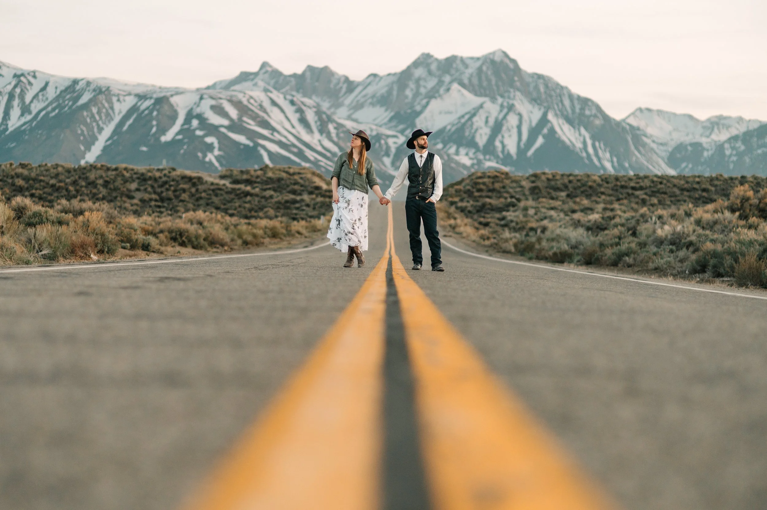 A man and woman holding hands standing in the middle of a mountain road, with snow-capped mountains in the background.