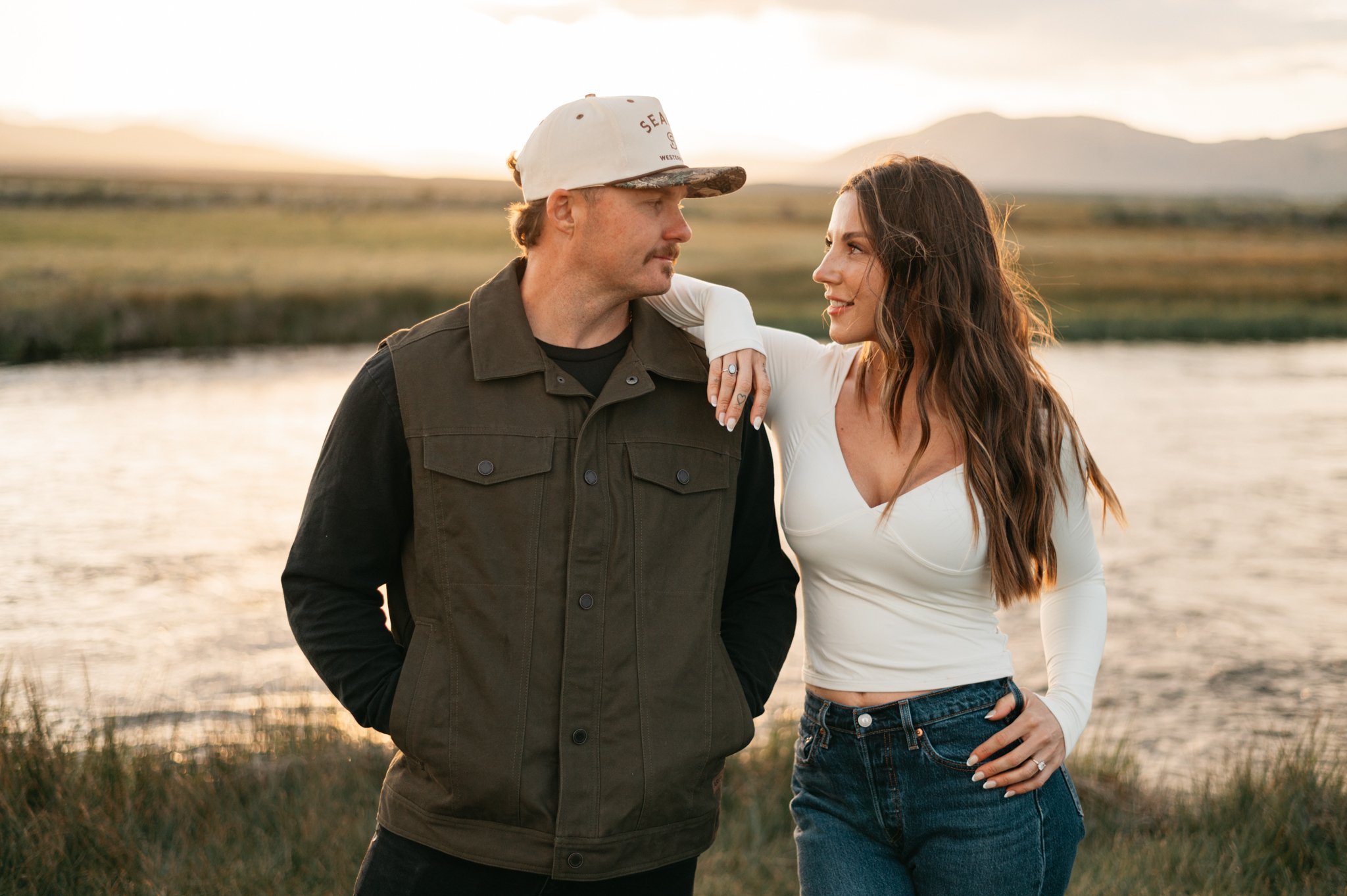 An engaged man and woman standing close by the owens river during sunset, looking at each other with the woman smiling. The man is wearing a baseball cap and a dark hunting vest, and the woman is in a white long-sleeve shirt and jeans, with her hand 