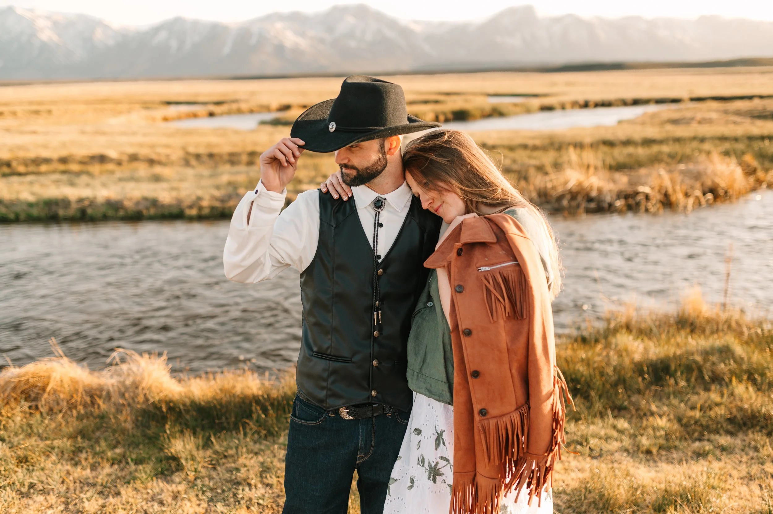 A man with a beard wearing a black cowboy hat and Western-style vest holding the brim of his hat, standing outdoors with a woman leaning her head on his shoulder. The woman has long hair and is wearing a brown fringe jacket and a white dress with flo
