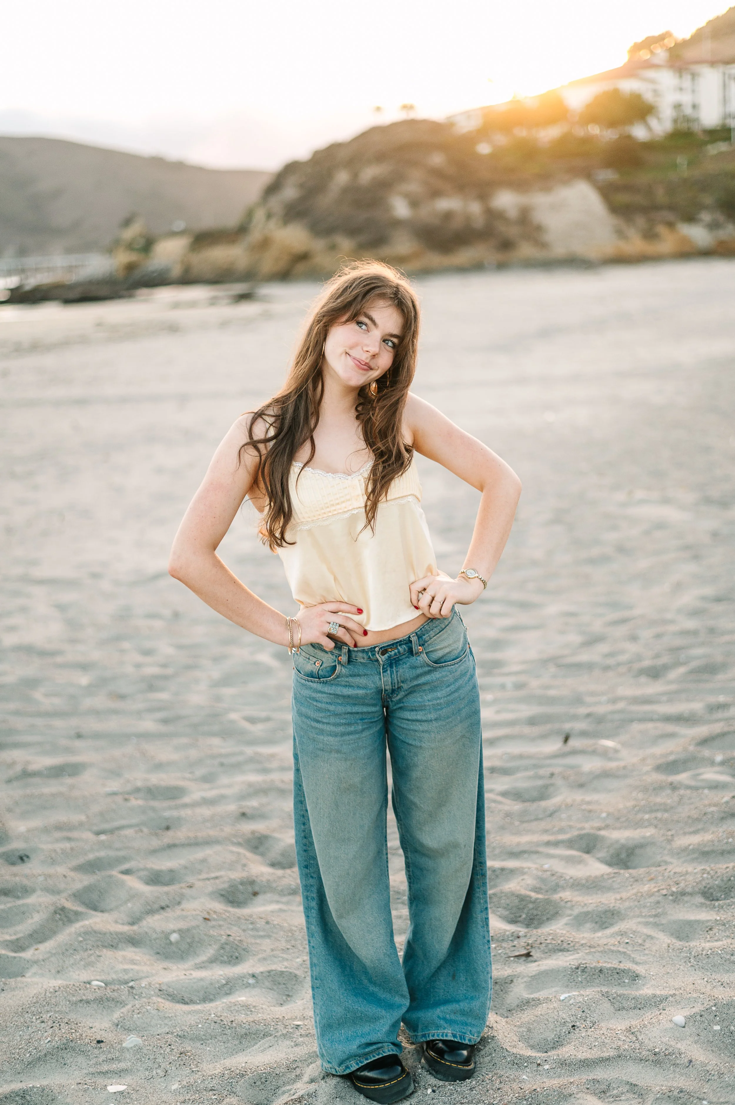A young woman with long brown hair standing on a sandy beach at sunset, wearing a yellow sleeveless top and wide-leg jeans, smiling with hands on hips.