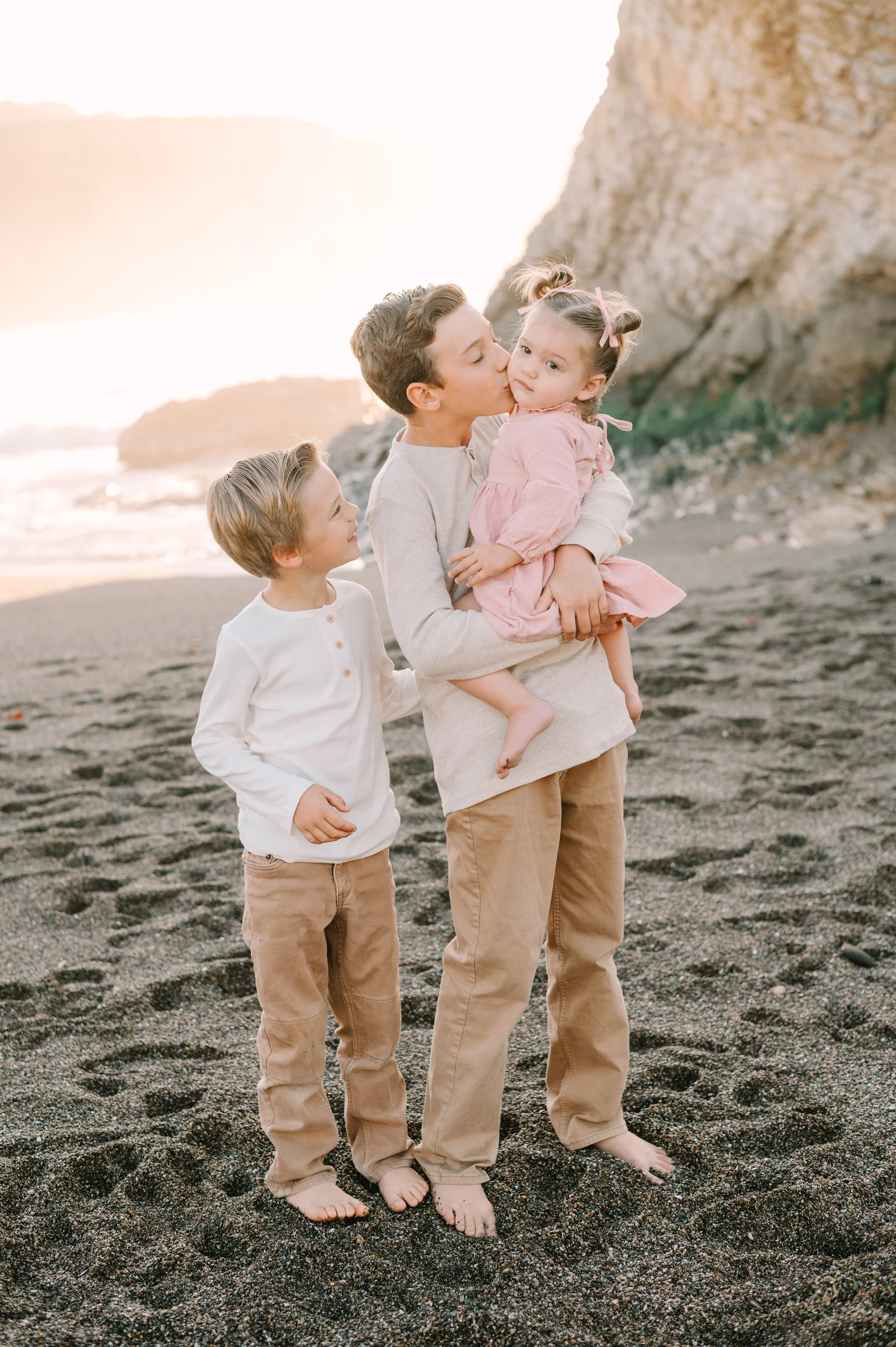 A family of four on a beach at sunset, with a rocky cliff in the background. The father is holding a young girl and giving her a kiss on the cheek while the mother and a young boy stand beside them, all barefoot on the sand.