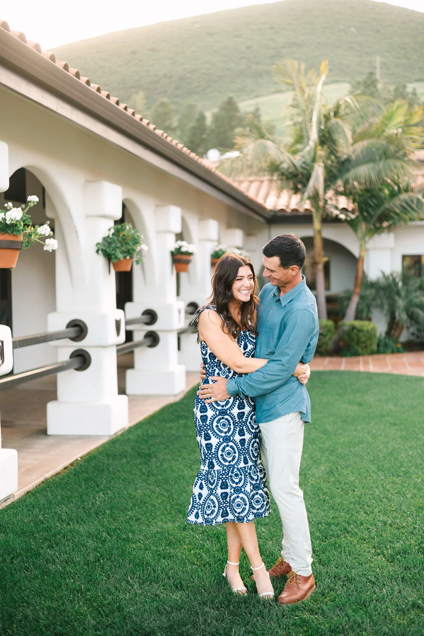 A happy couple embracing outdoors on a sunny day, with lush green grass and white building with arches and potted flowers in the background.