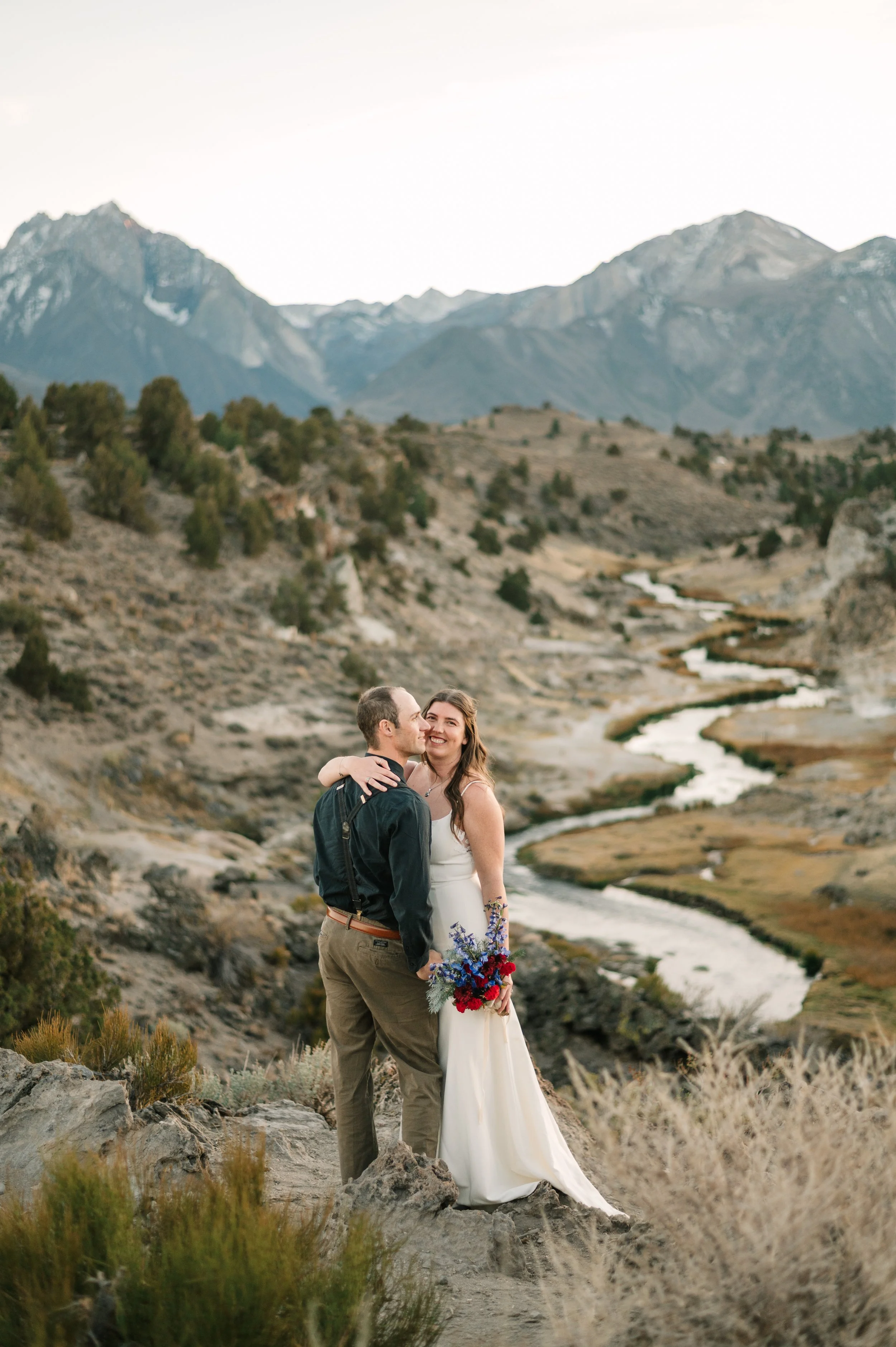 A couple dressed in wedding attire standing close together outdoors, smiling, with a mountain landscape and stream in the background.