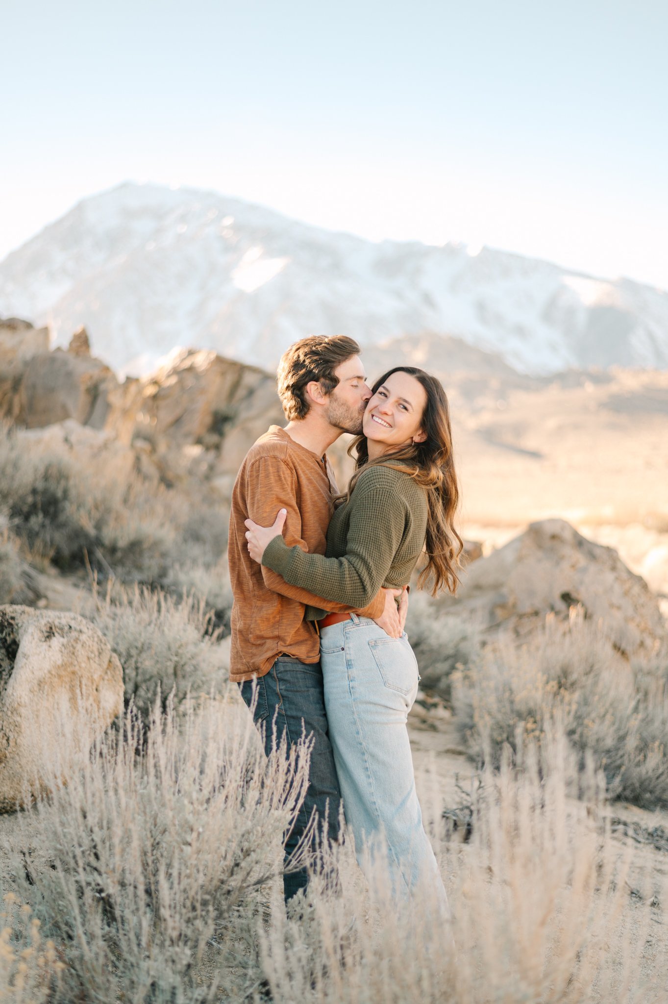 A couple hugging in a desert landscape with a mountain in the background, with the man kissing the woman's cheek.