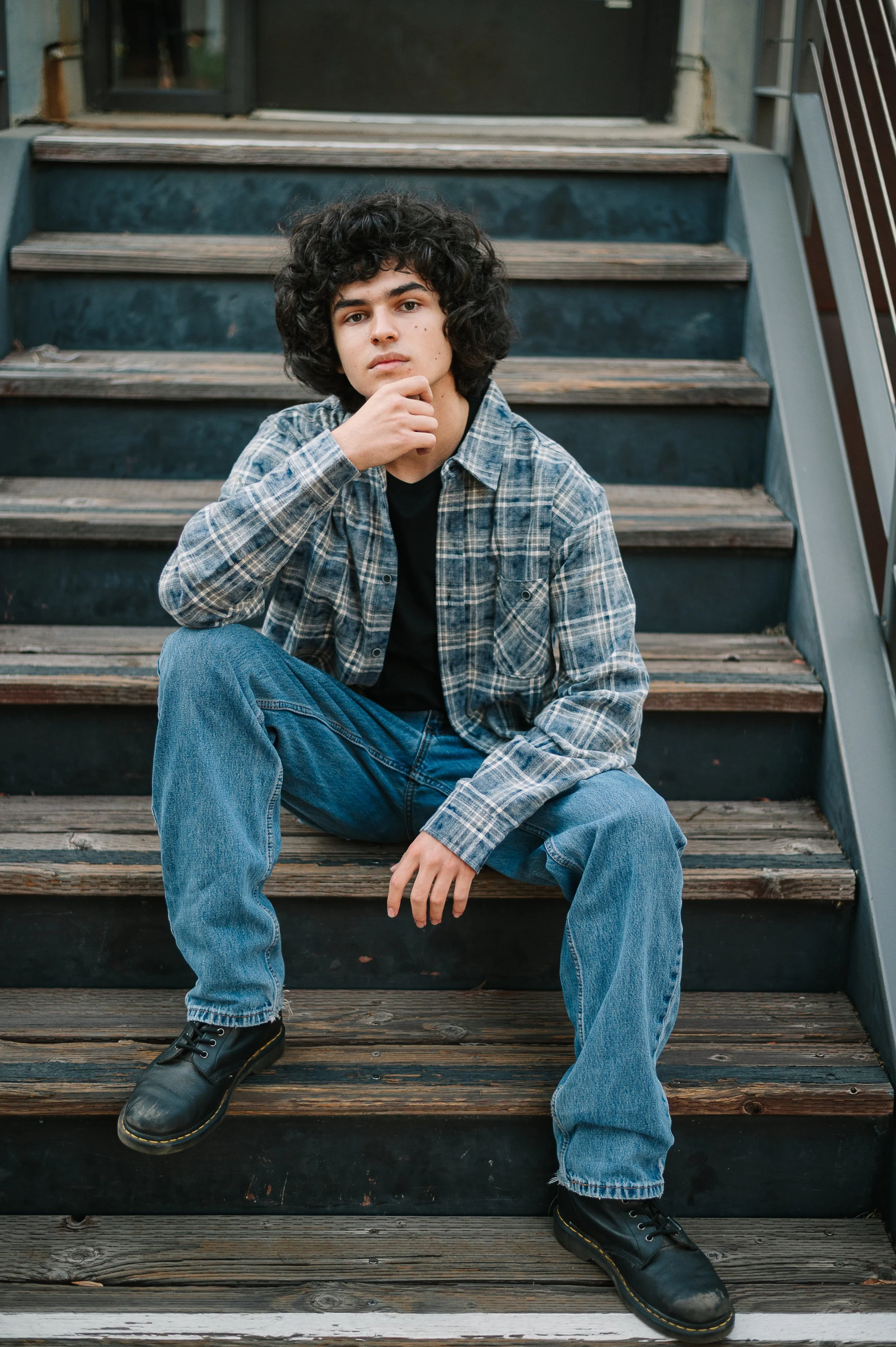 Young man sitting on a wooden staircase wearing a plaid shirt and jeans, with one hand on his chin and looking at the camera.