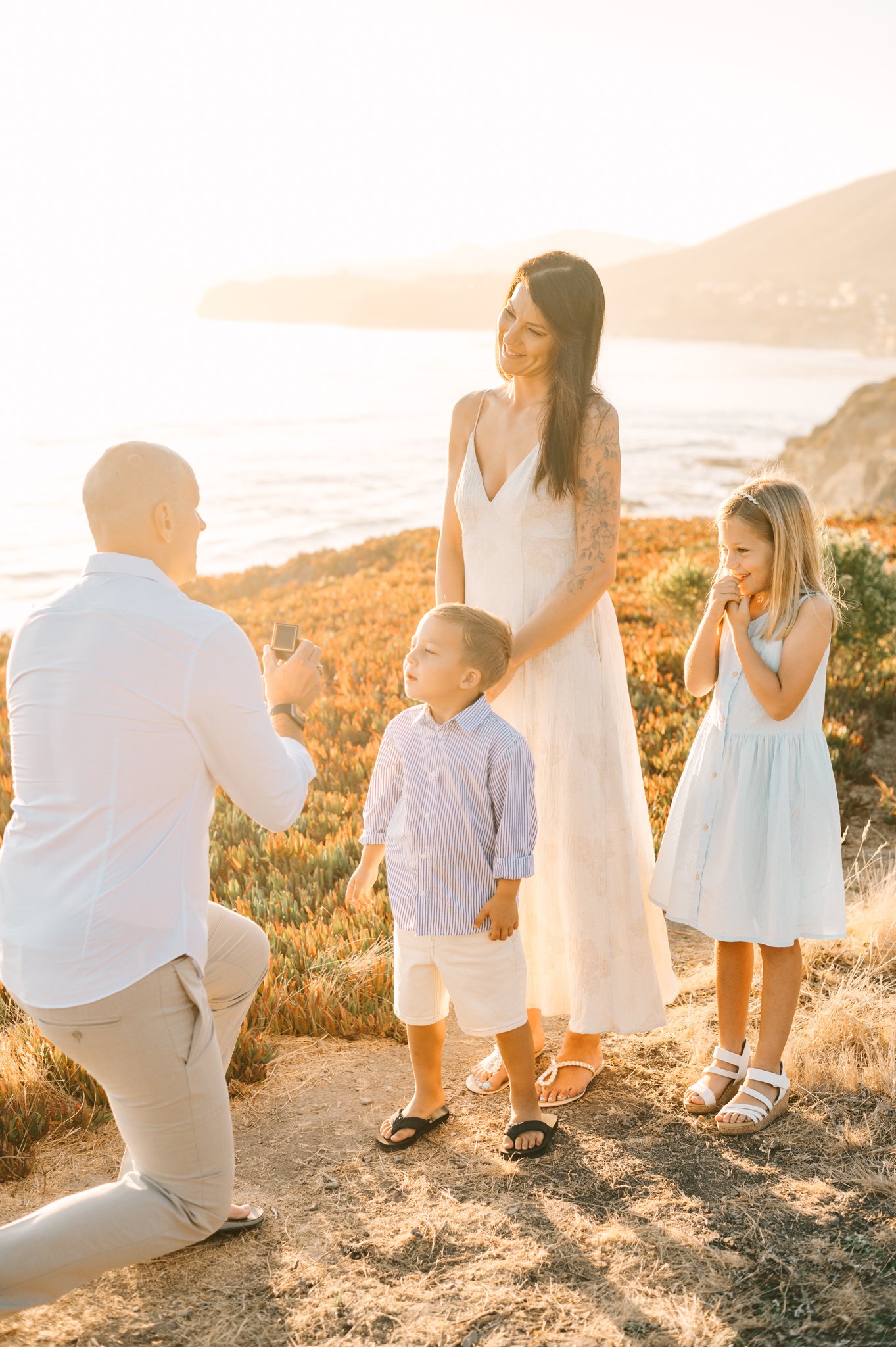 Family at sunset on a coastal hillside taking a photo together, including a woman, a young girl, a young boy, and a man kneeling with a camera.