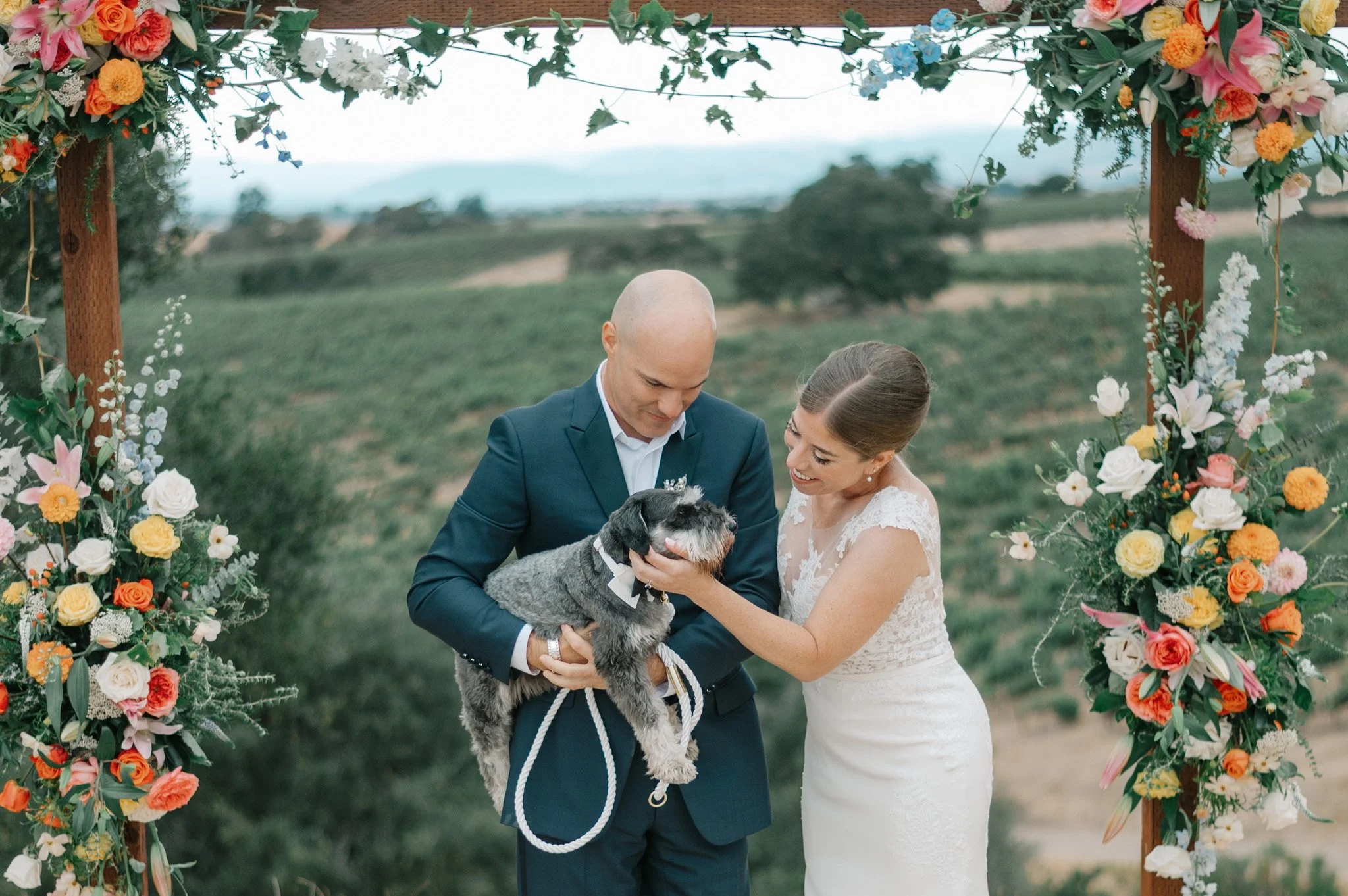 Bride and groom holding a dog under a floral wedding arch outdoors.