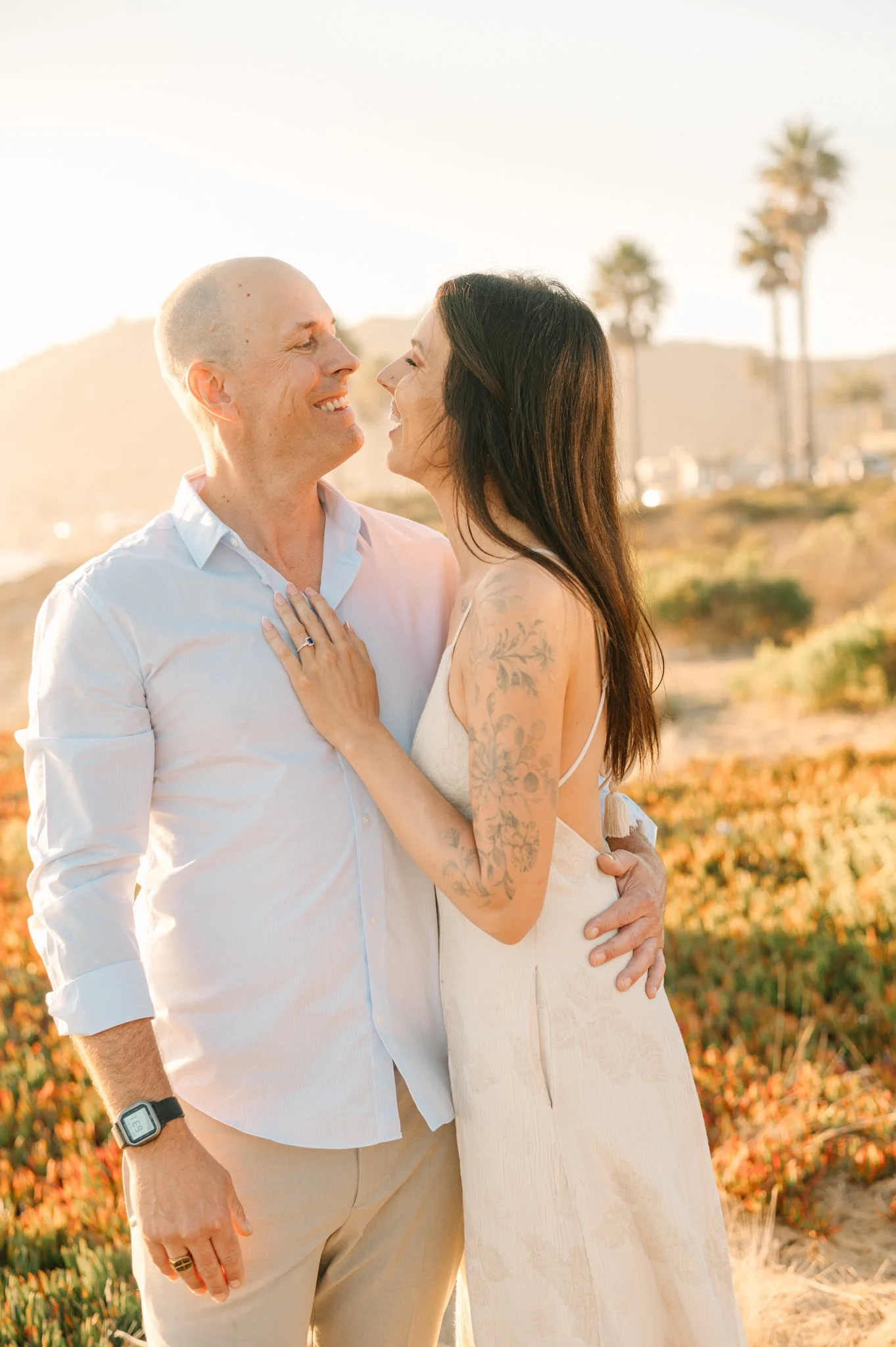 A couple smiling and looking at each other in a sunset outdoor setting with palm trees in the background.