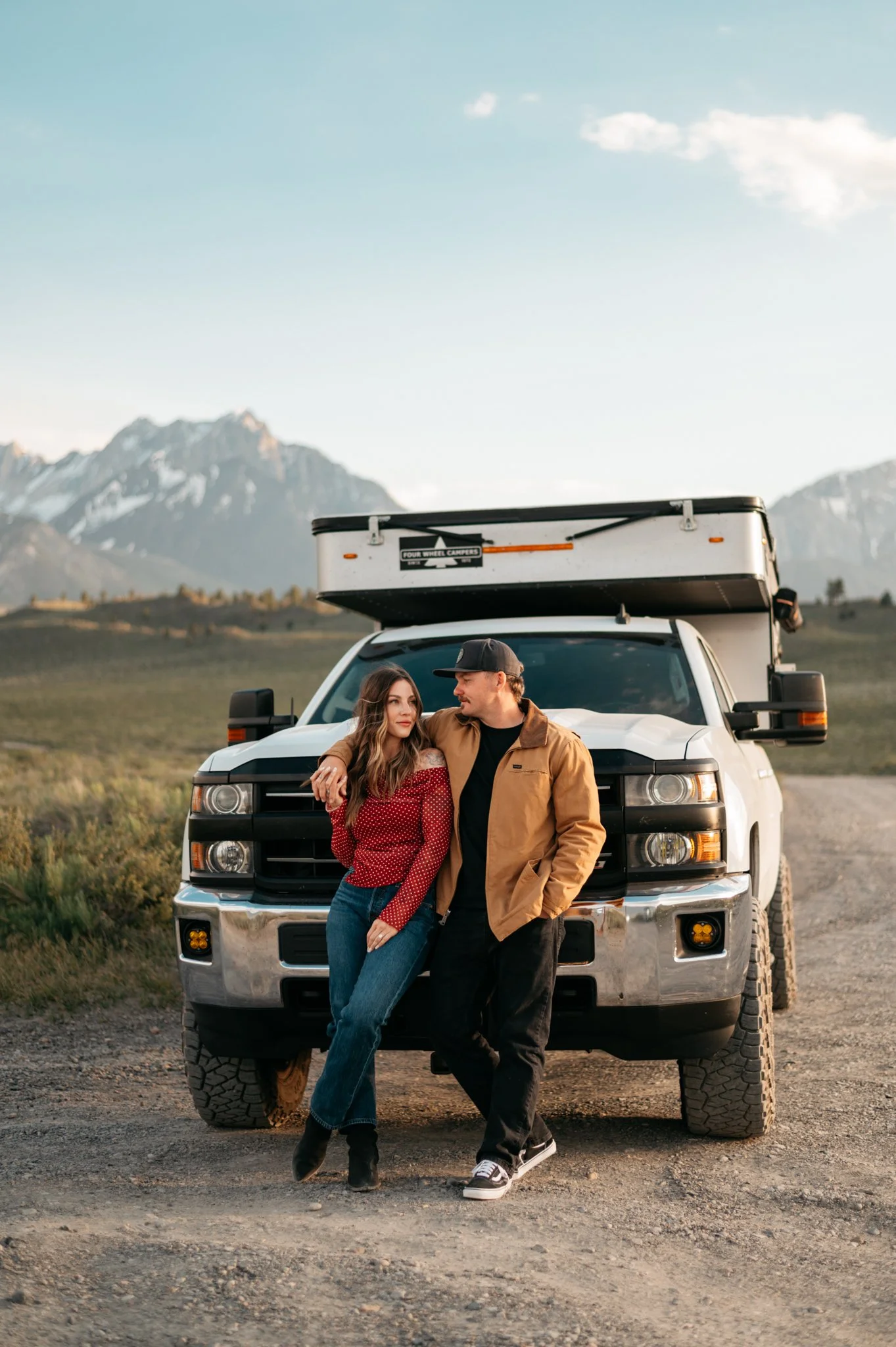 A young couple stands in front of a large white pickup truck with a mounted camper, on a dirt road in a rural mammoth mountain area during sunset for their engagement photos