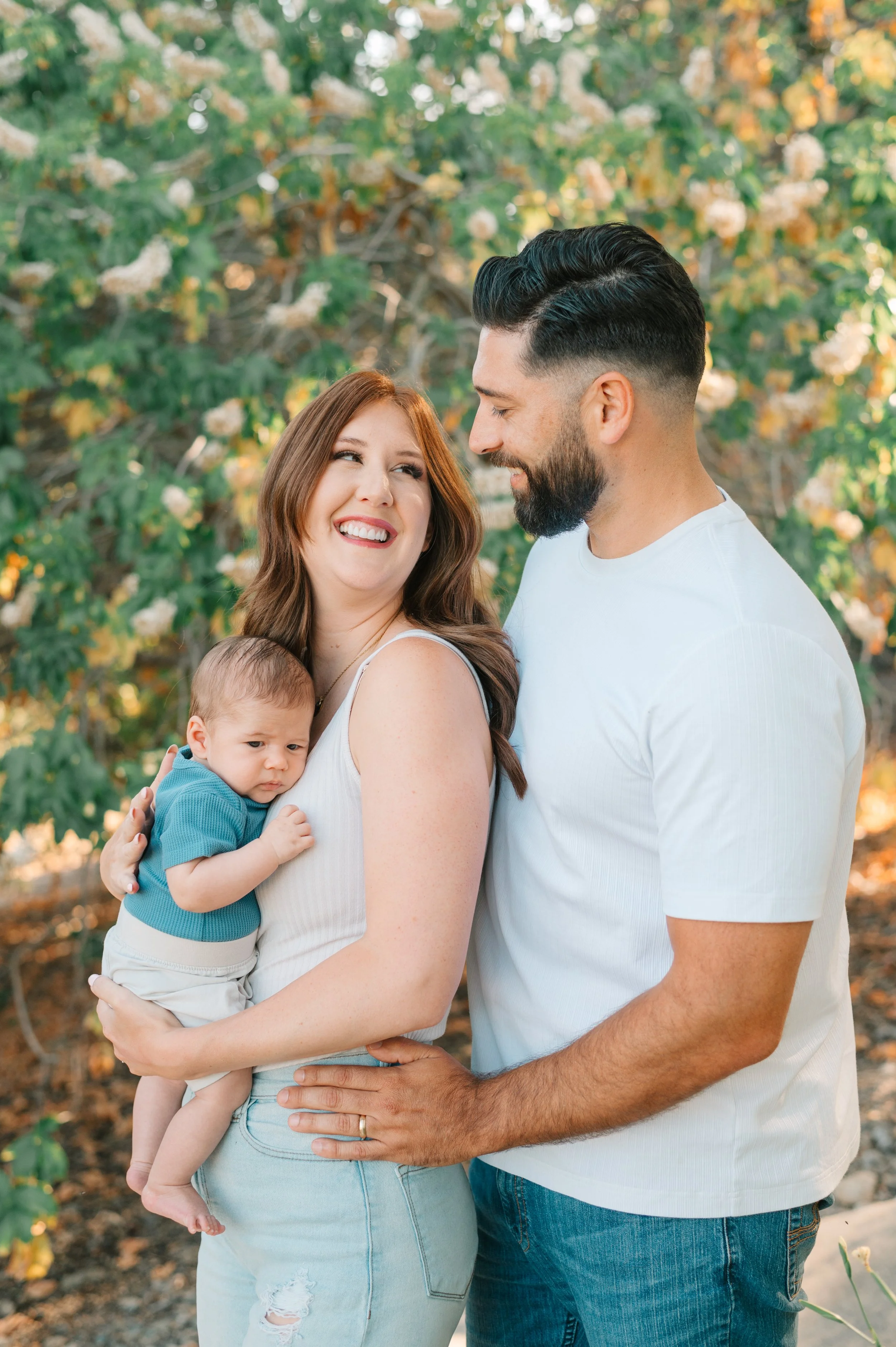 A happily smiling woman holding a baby, facing a man with a beard, outdoors with green foliage in the background.