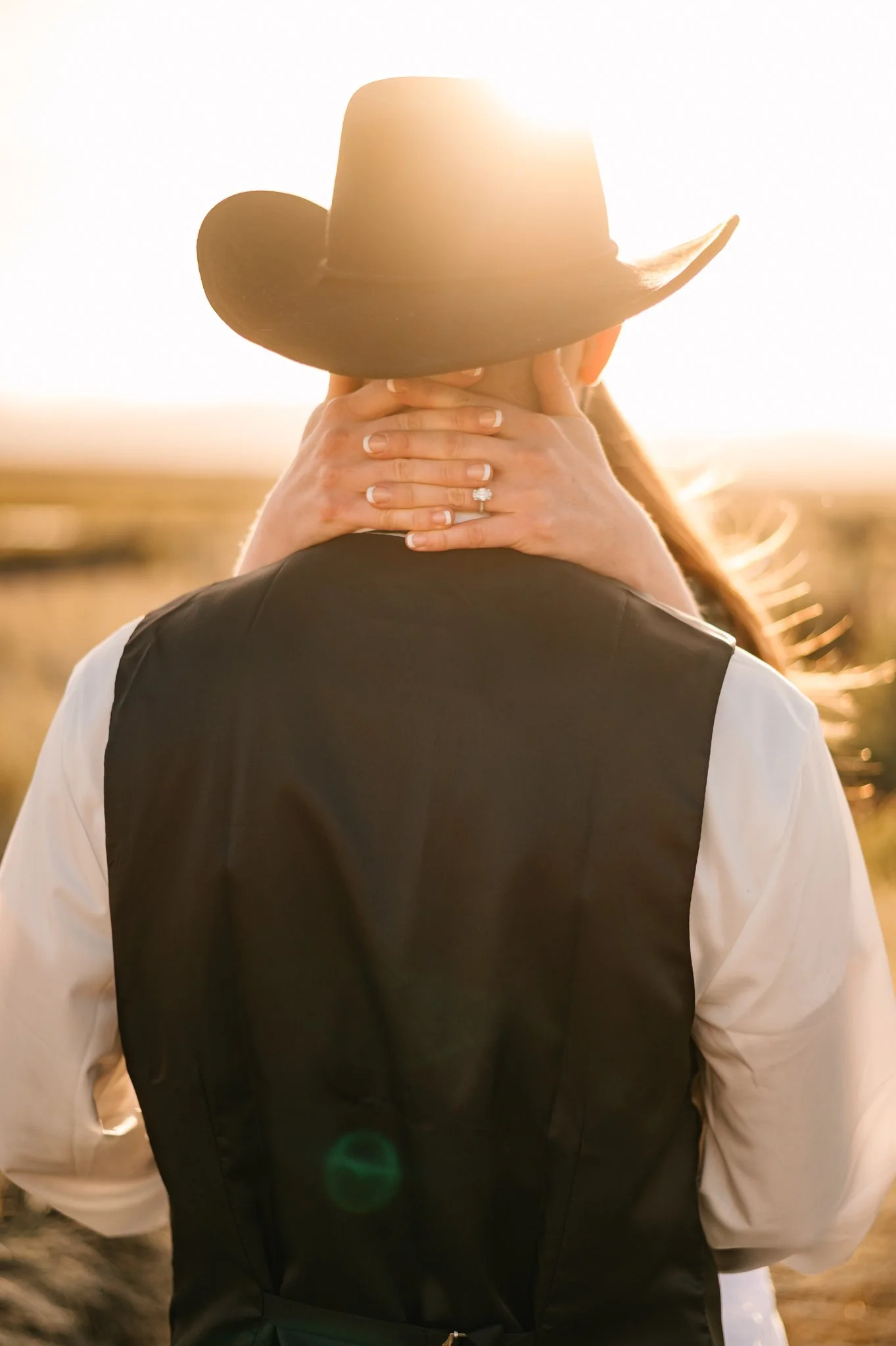 A person wearing a cowboy hat and a black vest, with hands resting on their neck with a big engagement ring, with sunlight bright behind them.