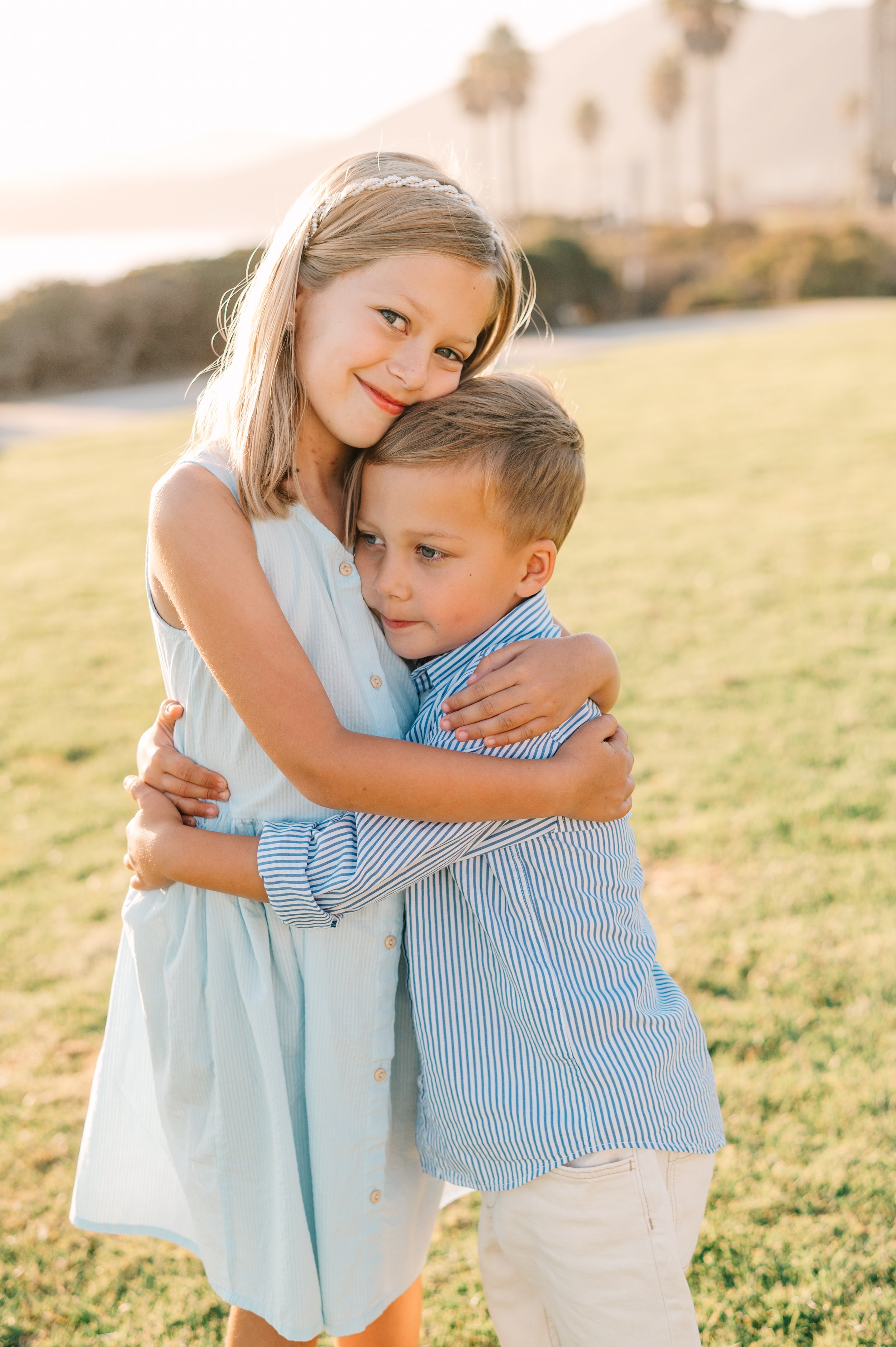 A girl with blonde hair wearing a white dress and a headband hugs a boy with light brown hair in a blue and white striped shirt outdoors on a sunny day.