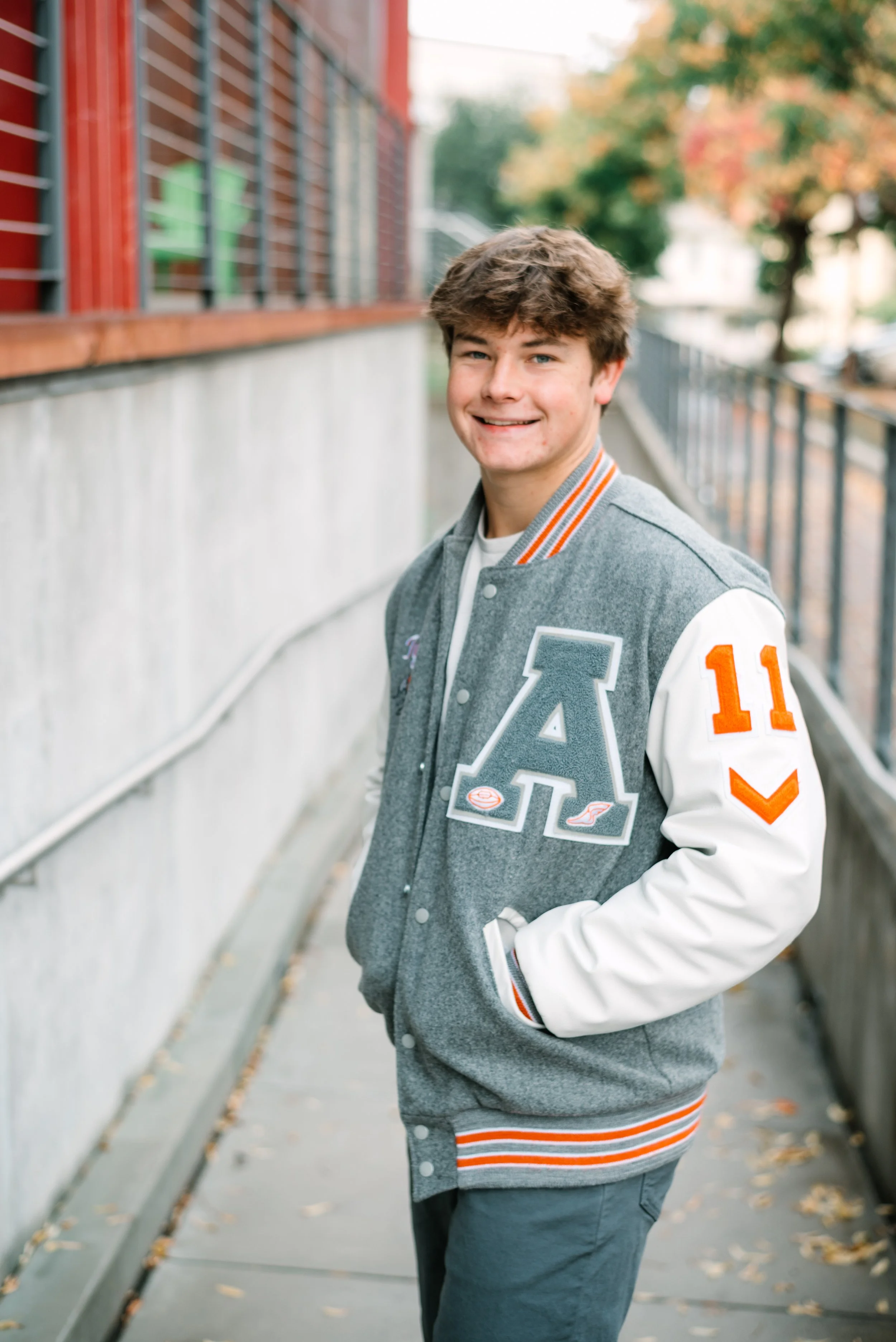 A young man with brown hair standing outdoors on a sidewalk, smiling and wearing a gray varsity jacket with white sleeves, orange accents, and the letter 'A' on the front.
