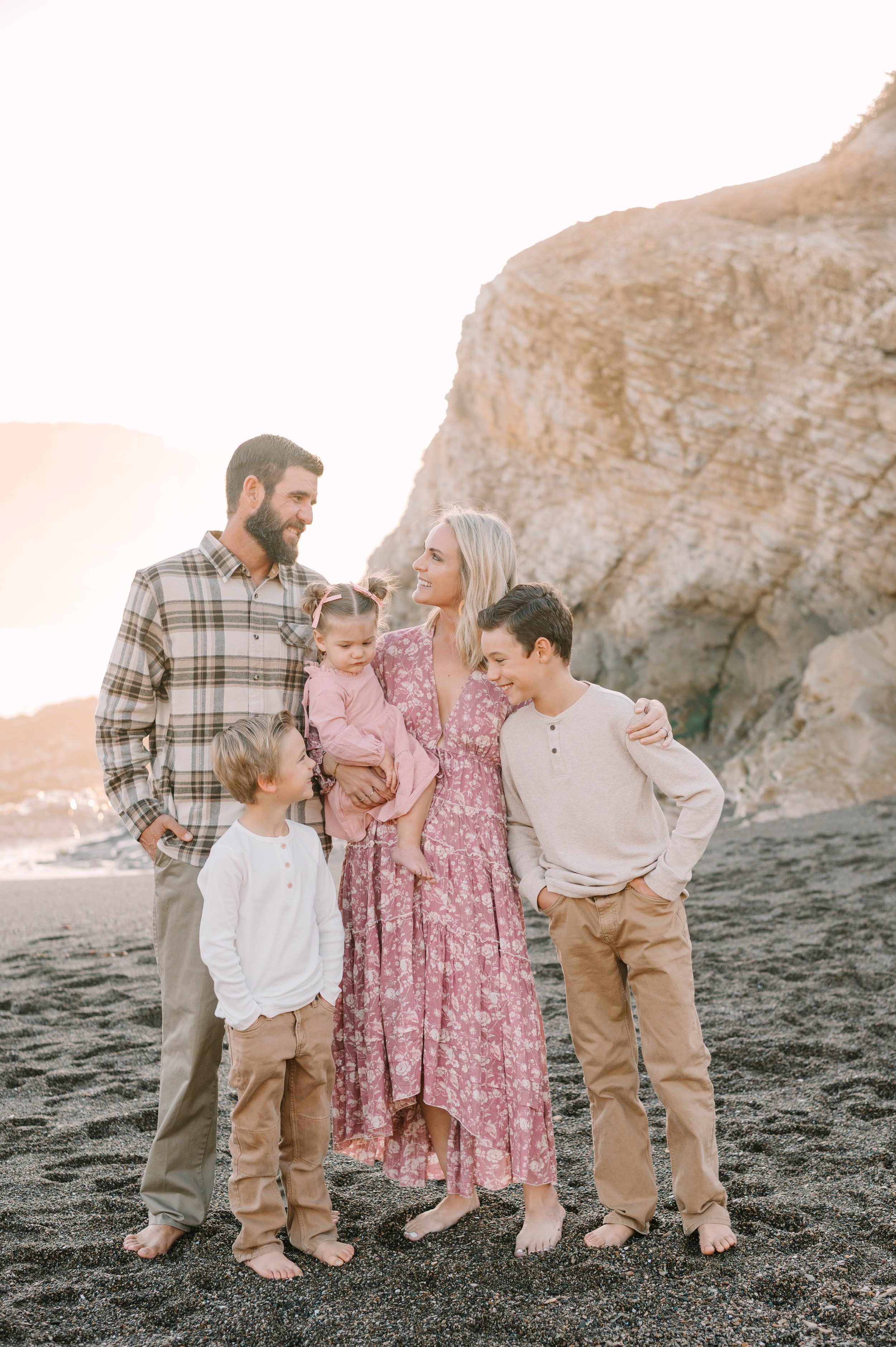 A family of six standing barefoot on a black sand beach with rocky cliffs in the background during sunset, smiling and looking at each other.