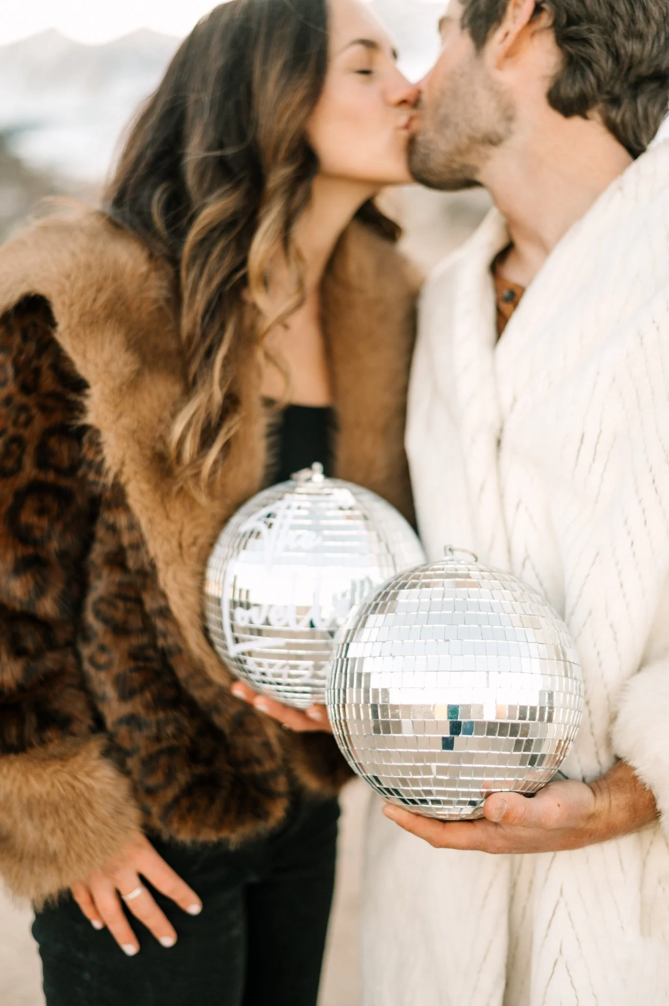 A couple kissing while holding disco balls, one with a word 'fever' visible, on a beach or outdoor setting.
