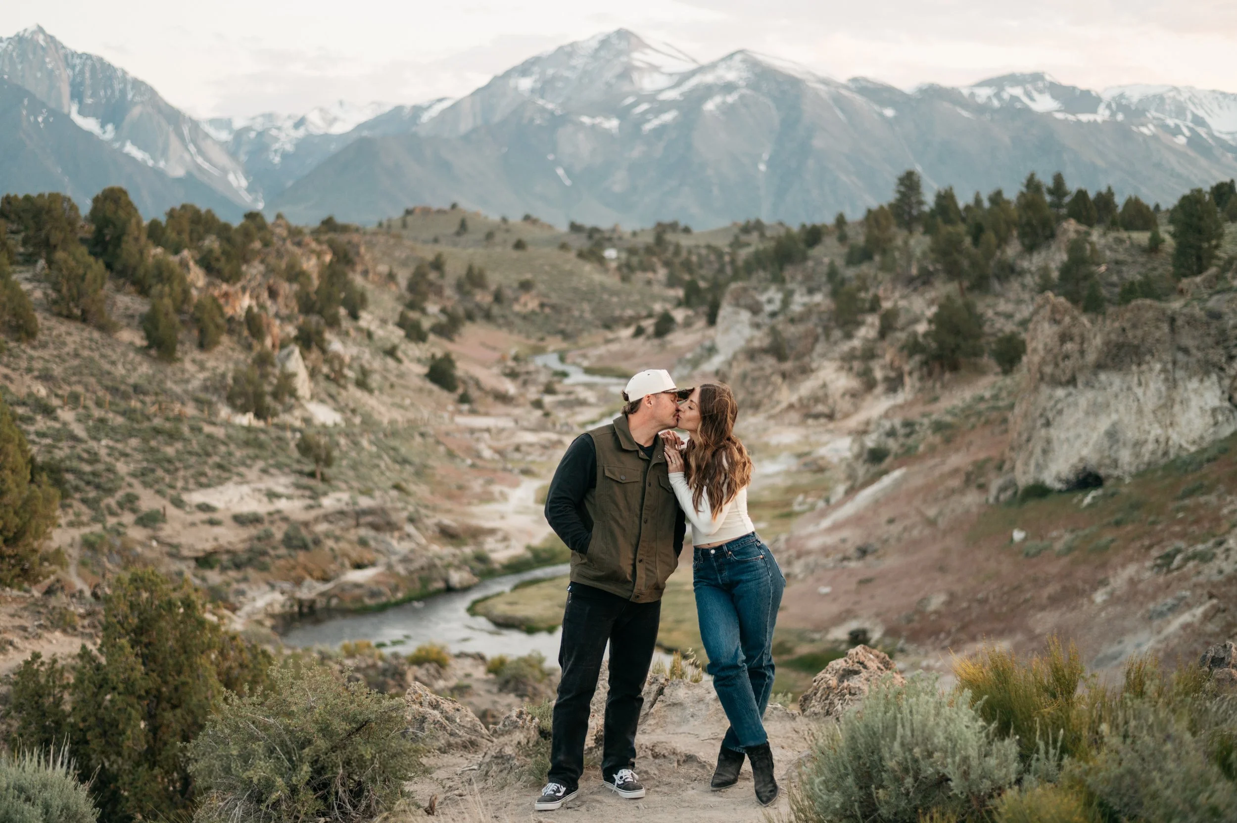 A young couple shares a kiss in a mountainous outdoor setting with snow-capped peaks in the background, a river flowing through the valley, and sparse vegetation.