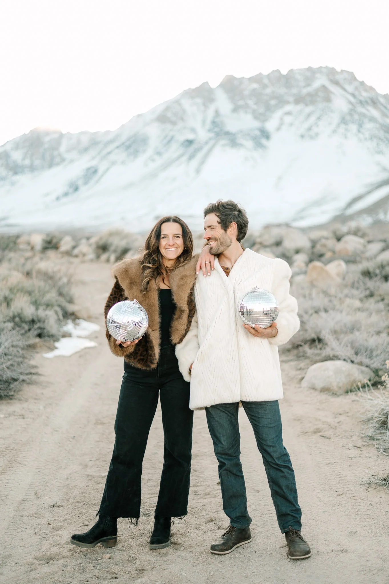 A happy couple standing on a dirt path in front of a snow-capped mountain, holding disco balls, dressed in winter clothes.