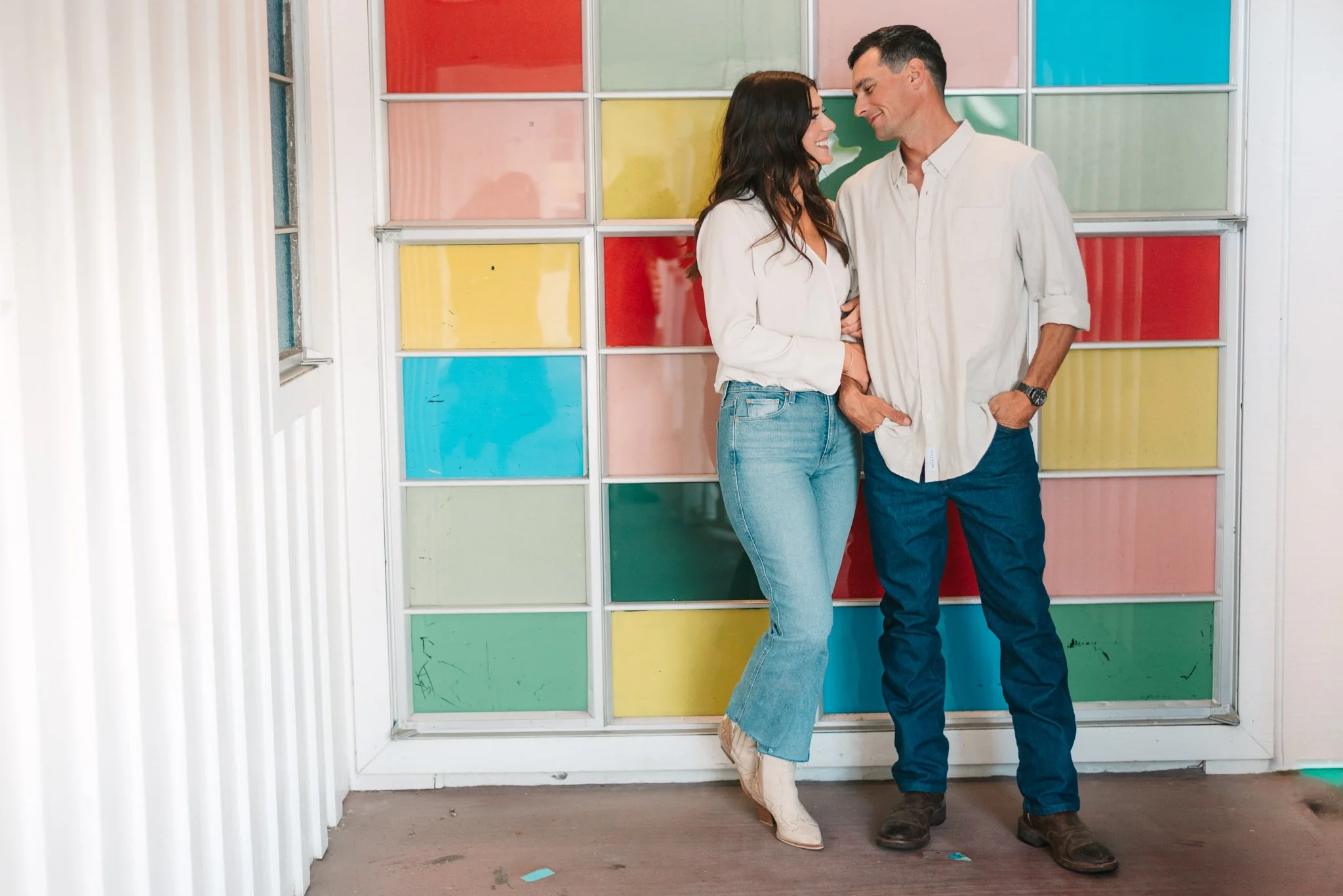 A man and woman standing close together, smiling and looking at each other, against a colorful, stained-glass style window with red, yellow, green, blue, and pink sections. The woman is wearing a white blouse and light blue jeans, and the man is wear
