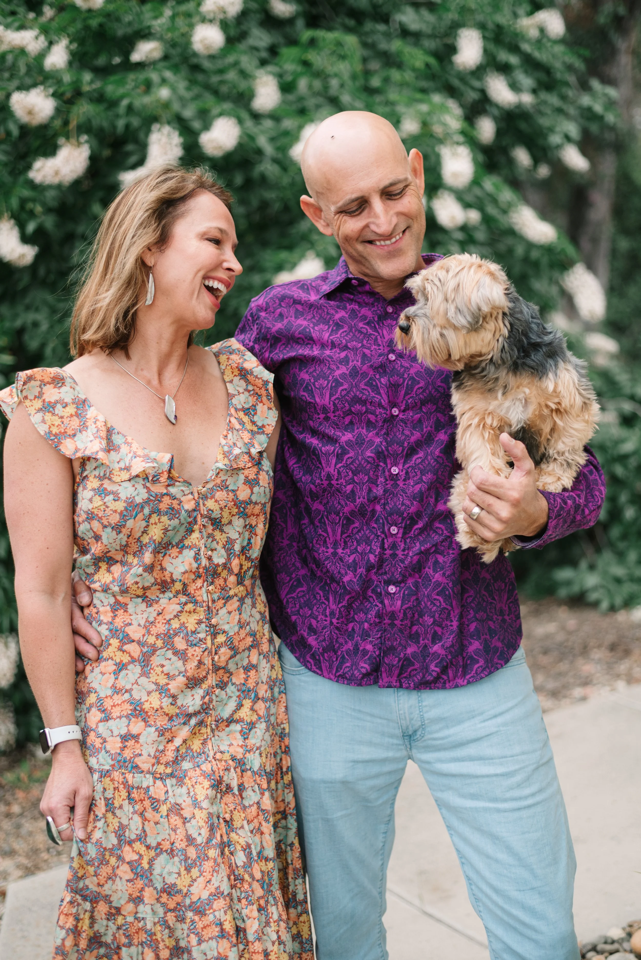 A smiling woman in a floral dress standing next to a smiling man holding a small dog outdoors in a garden with white flowers.