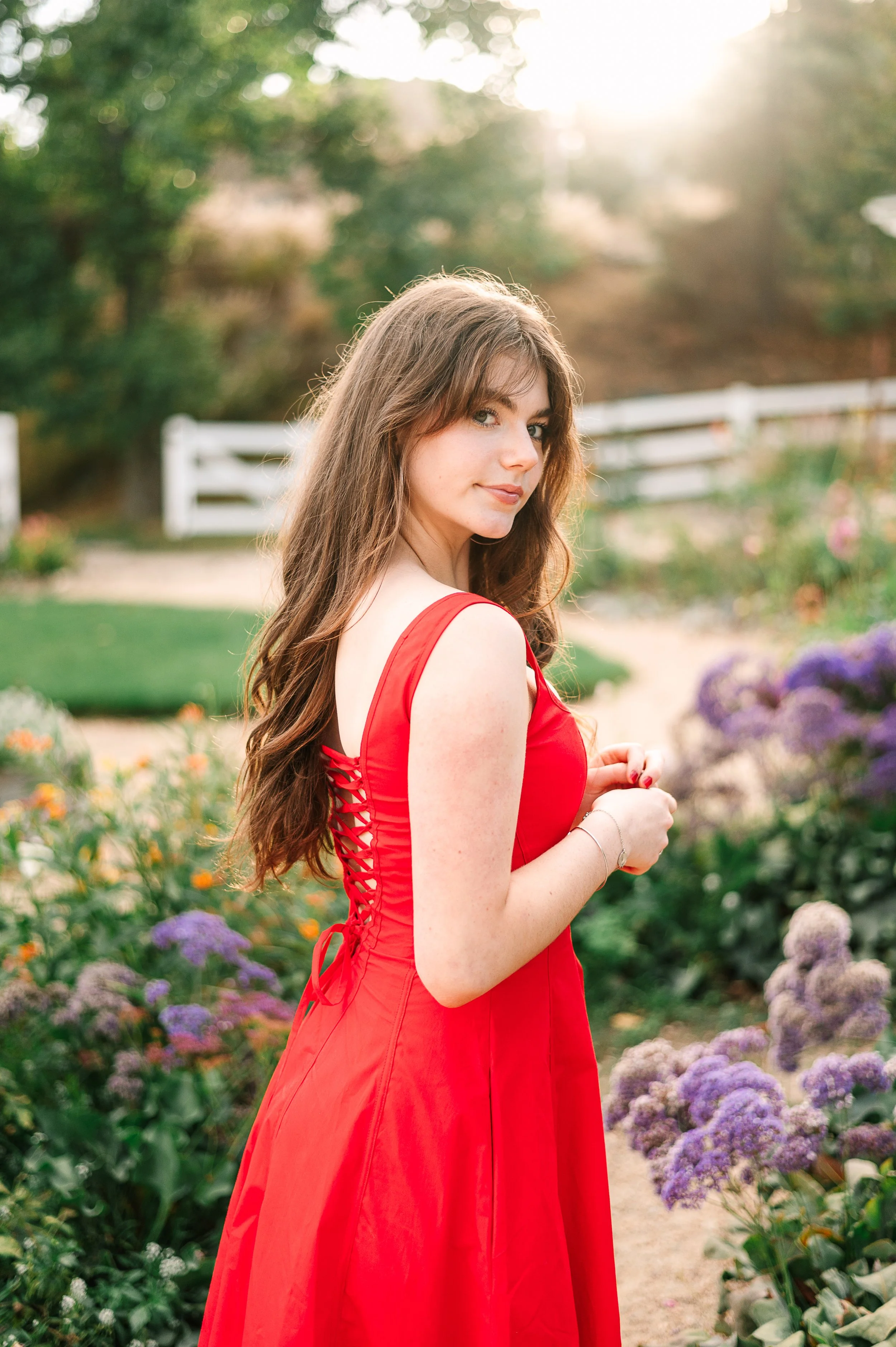Young woman with brown hair in a red dress standing in a garden with purple flowers, white fence, and trees in the background, illuminated by sunlight.