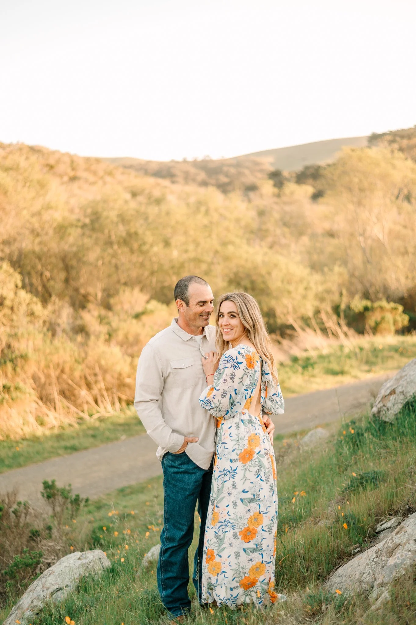 A couple standing close together outdoors in a scenic natural setting during golden hour, smiling at each other. The man is wearing a light-colored shirt and jeans, and the woman is dressed in a floral maxi dress.