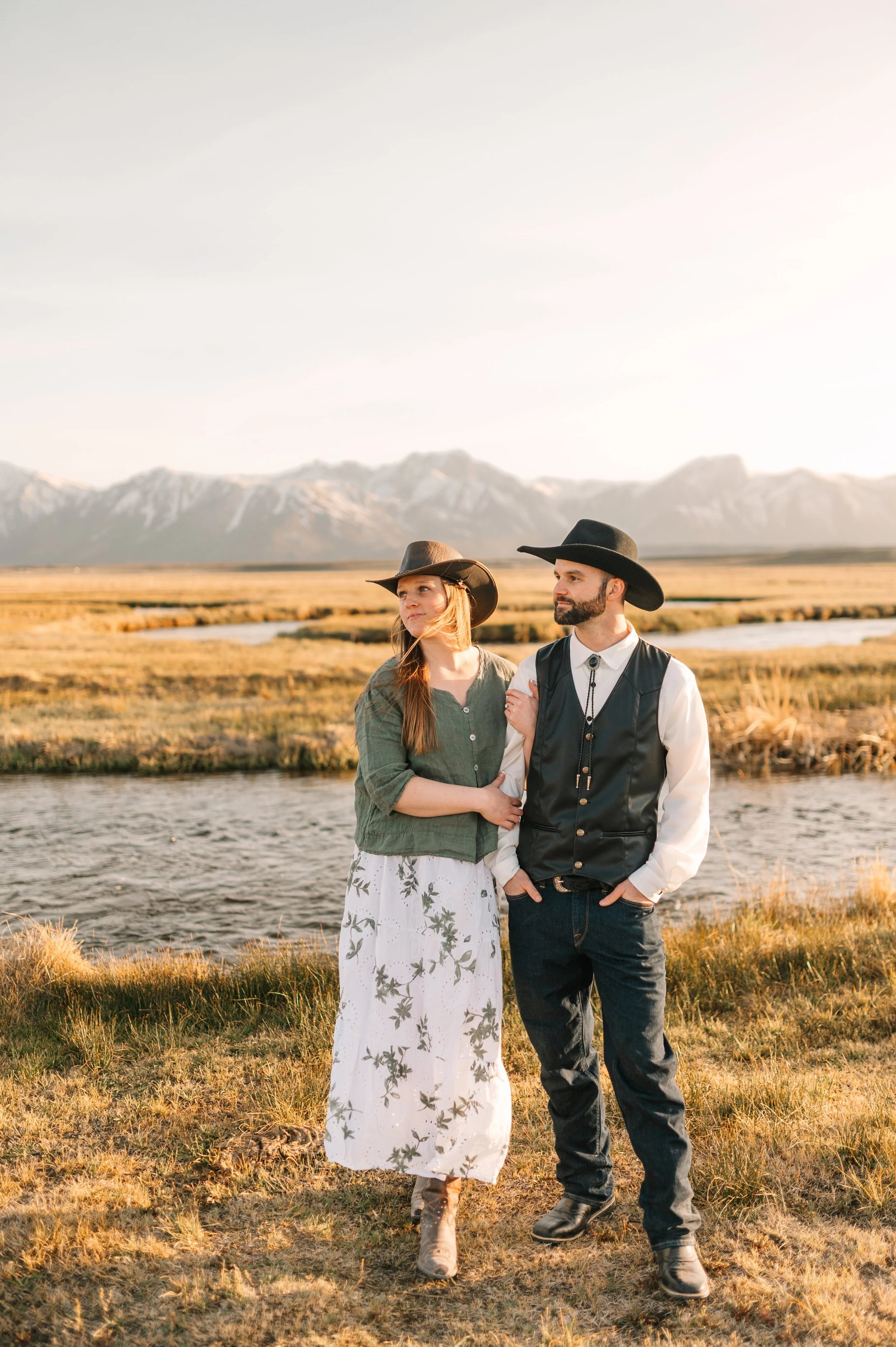 A couple standing outdoors by a river with a mountain range in the background, dressed in western-style clothing, with the woman in a long white floral skirt and brown hat, and the man in dark jeans, a vest, and a black cowboy hat, during sunset.