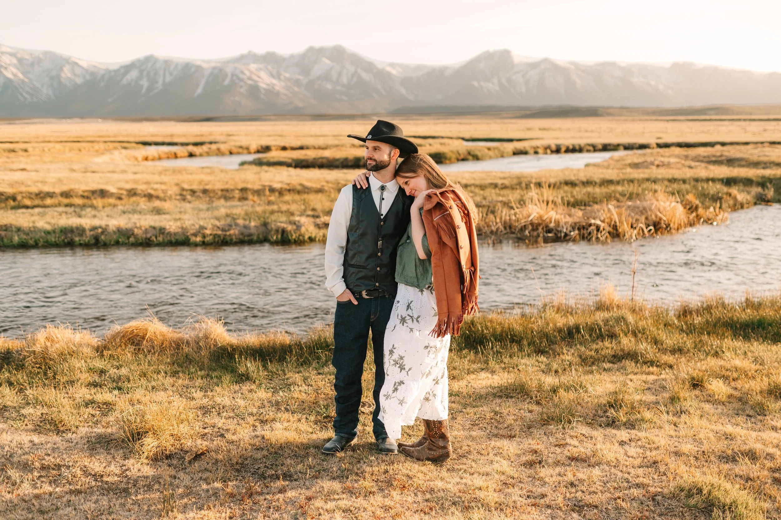A man and woman standing by a river in a rural landscape with mountains in the background. The man is wearing a cowboy hat and vest, and the woman is leaning her head on his shoulder wearing a patterned skirt and a jacket.