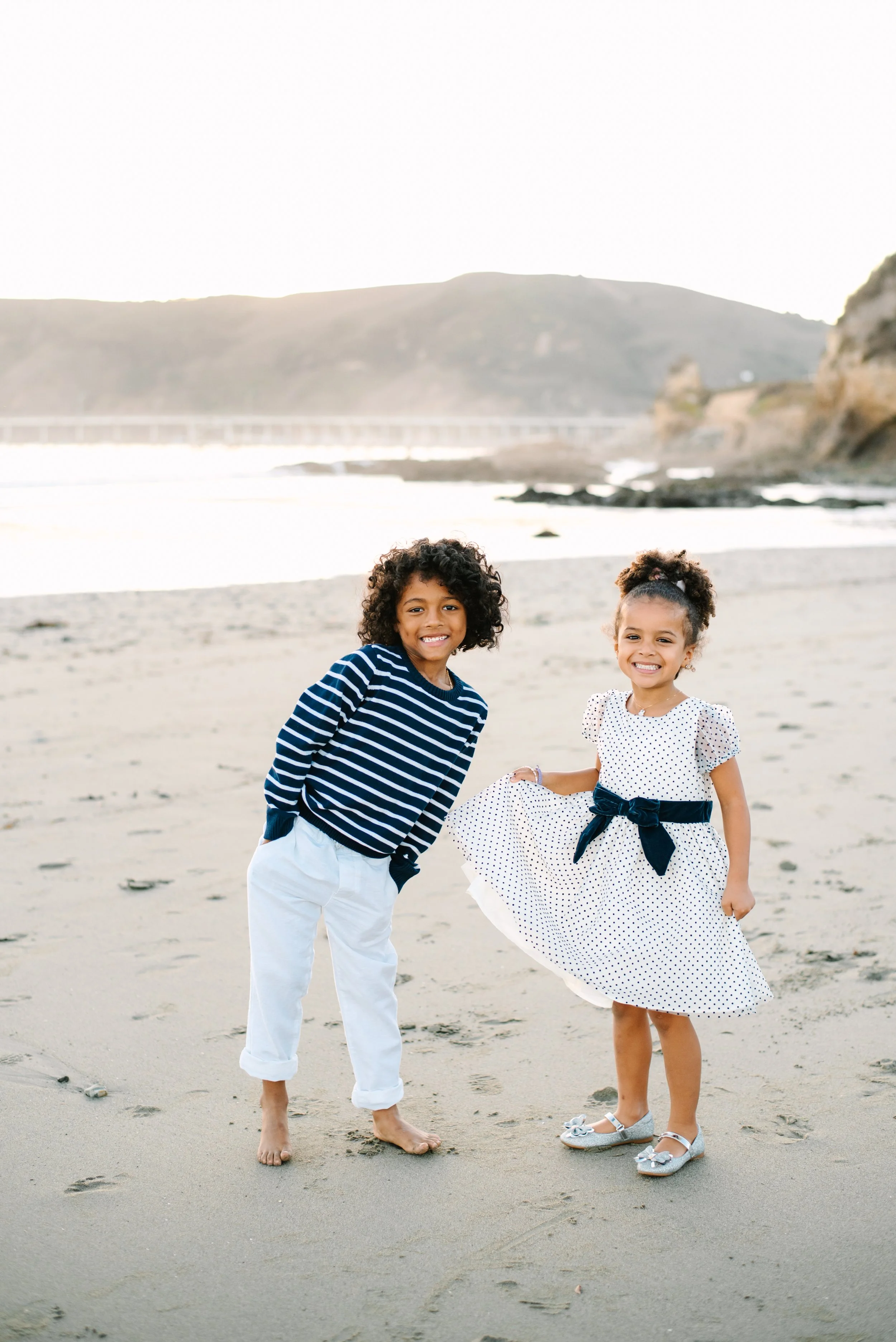 Two young children, a boy and a girl, standing barefoot on a sandy beach near the ocean, smiling at the camera with a coastal landscape in the background.