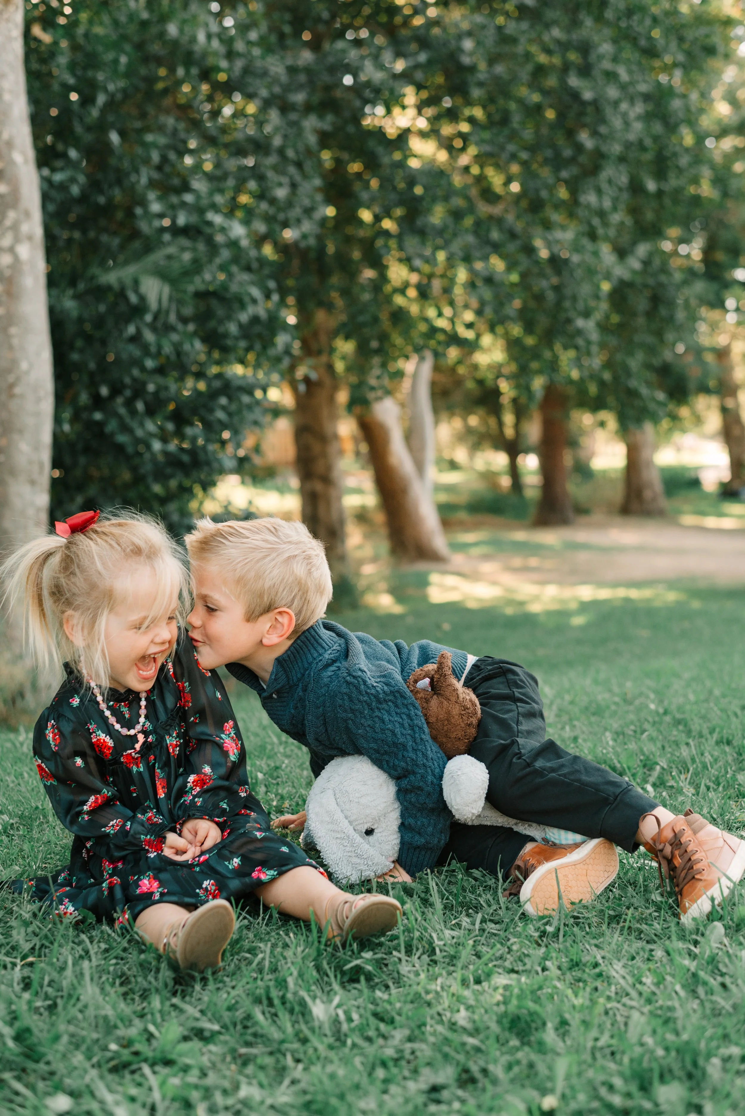 Two children, a girl and a boy, sitting on the grass in a park, playing and whispering to each other with trees in the background.