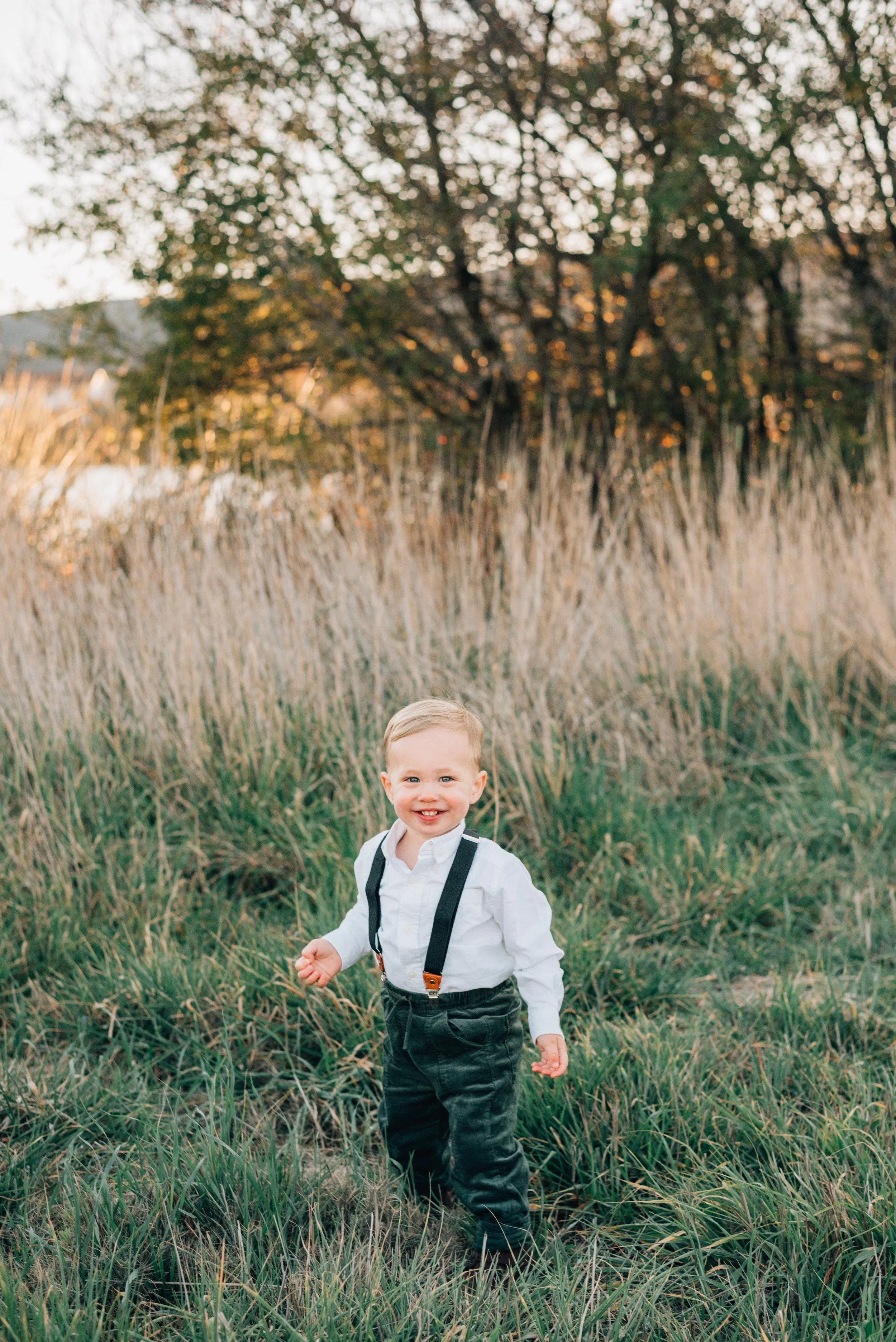 A young boy smiling and standing in a grassy field with tall grasses and trees in the background during the daytime.