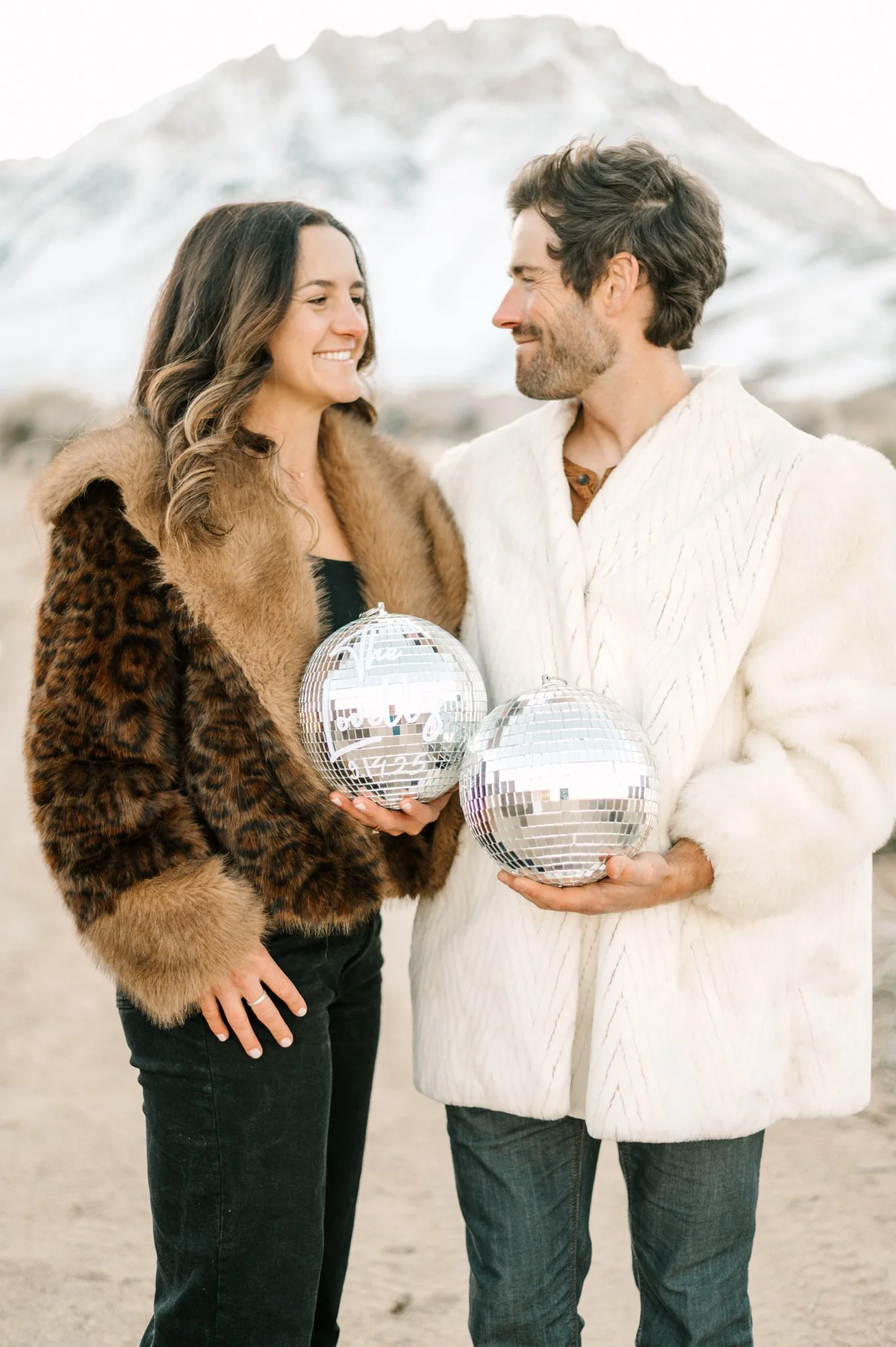 A woman and man are standing outdoors in a snowy landscape, smiling and looking at each other. The woman is wearing a fur coat, and the man is in a white knit sweater. They are holding disco balls with written messages and numbers on them.
