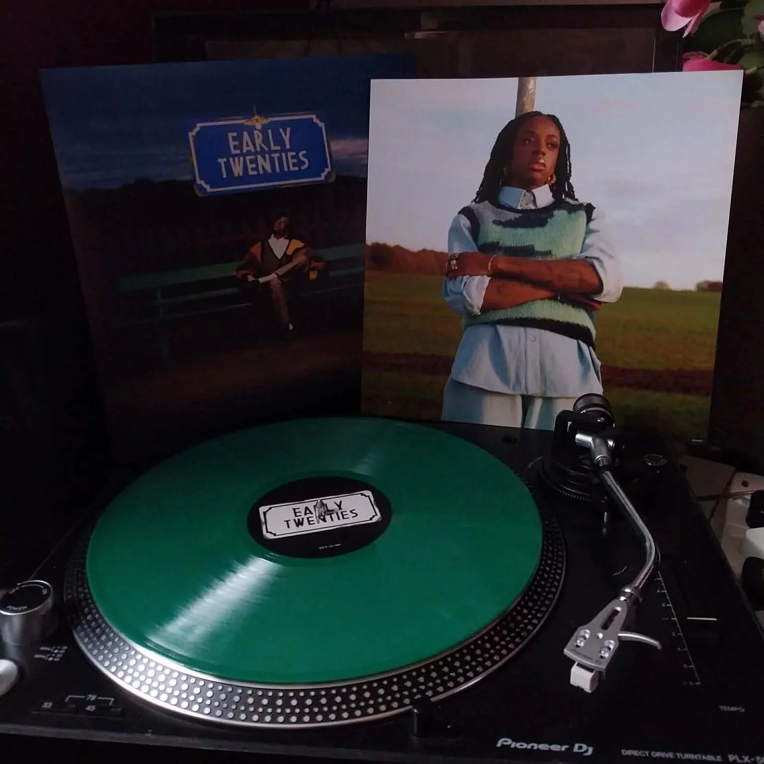 a deep green vinyl disc on a Pioneer turntable, speakers at either side. Stood behind is the vinyl sleeve for ‘Early Twenties’ by Cat Burns, which features a photo of her on the front sat at a train station.