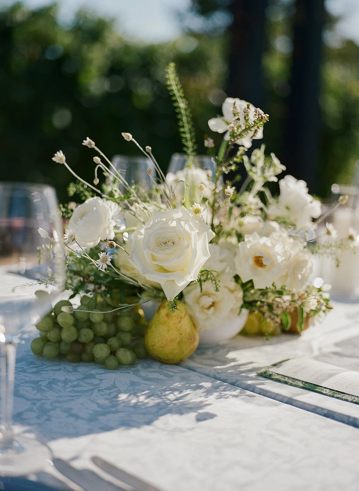 A floral centerpiece with white roses, small white flowers, a pear, and green grapes on a white tablecloth, with sunlight and blurred green background.
