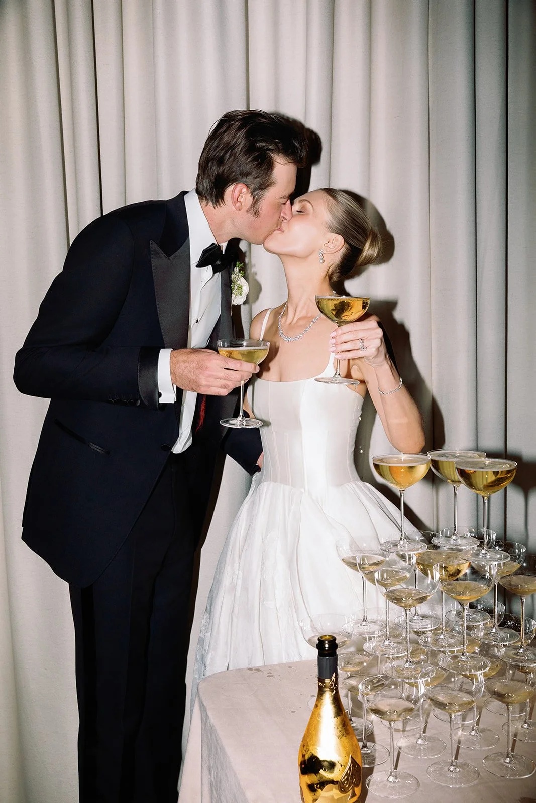 A newlywed couple is kissing at their wedding reception, holding champagne glasses. The groom is wearing a black tuxedo and the bride is in a white wedding dress. There are several champagne glasses and a champagne bottle on the table nearby.