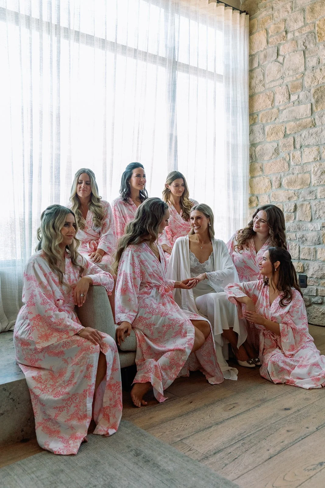 Bridal party of nine women, some sitting and some standing, sharing a moment together in a room with a large window with sheer curtains and a stone wall.