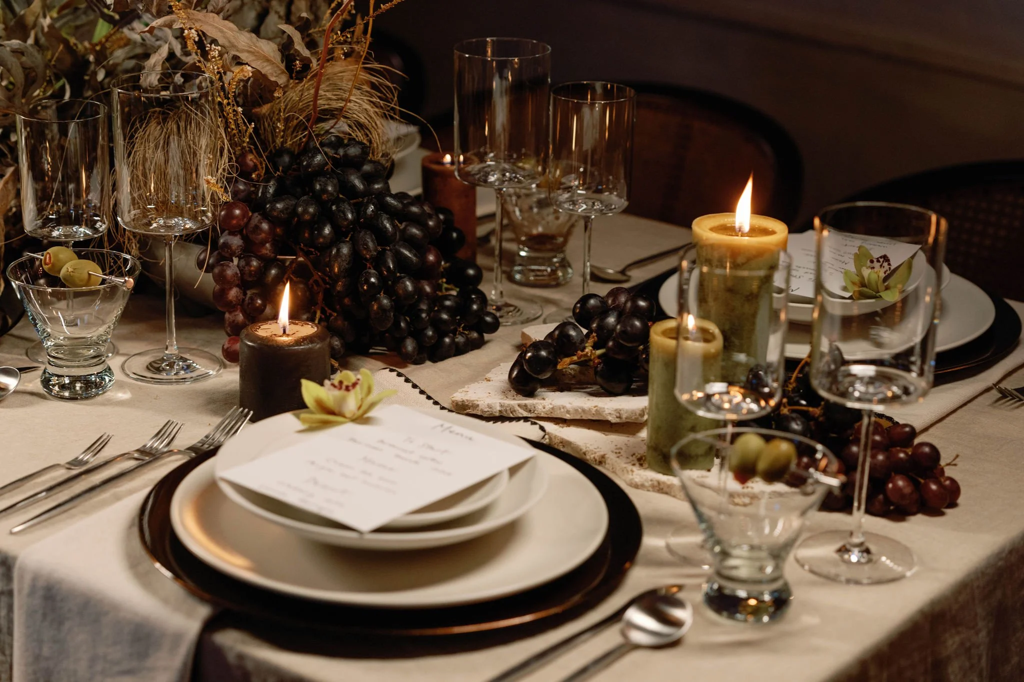 Elegant dining table set for a formal dinner with a beige tablecloth, multiple wine glasses, a menu on a plate, black and green grapes, and lit candles creating a warm ambiance.