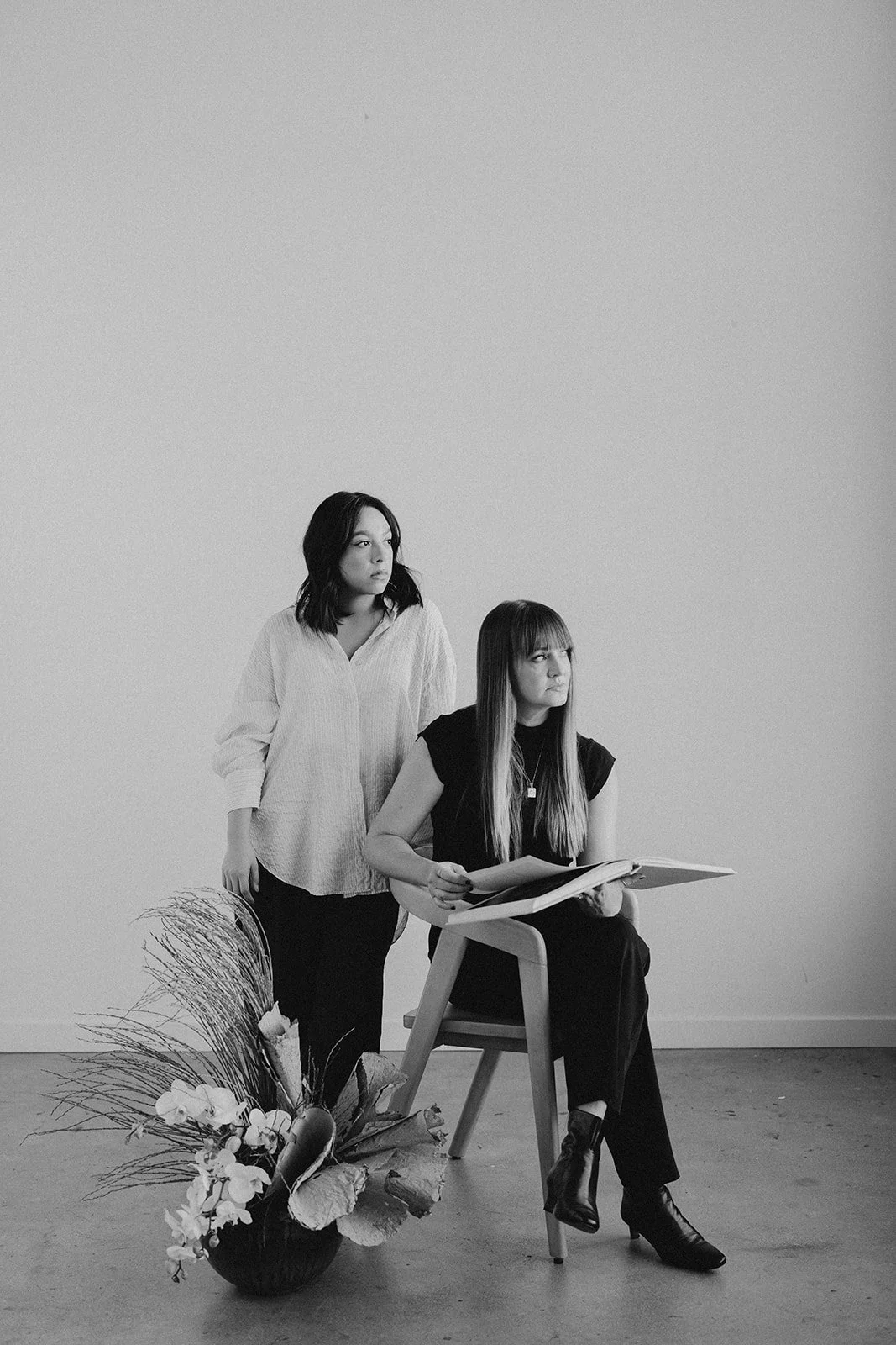 Black and white photo of two women in a minimalist room. One woman is seated on a chair, holding an open book, and the other woman is standing beside her. There is a large flower arrangement on the floor to the left.